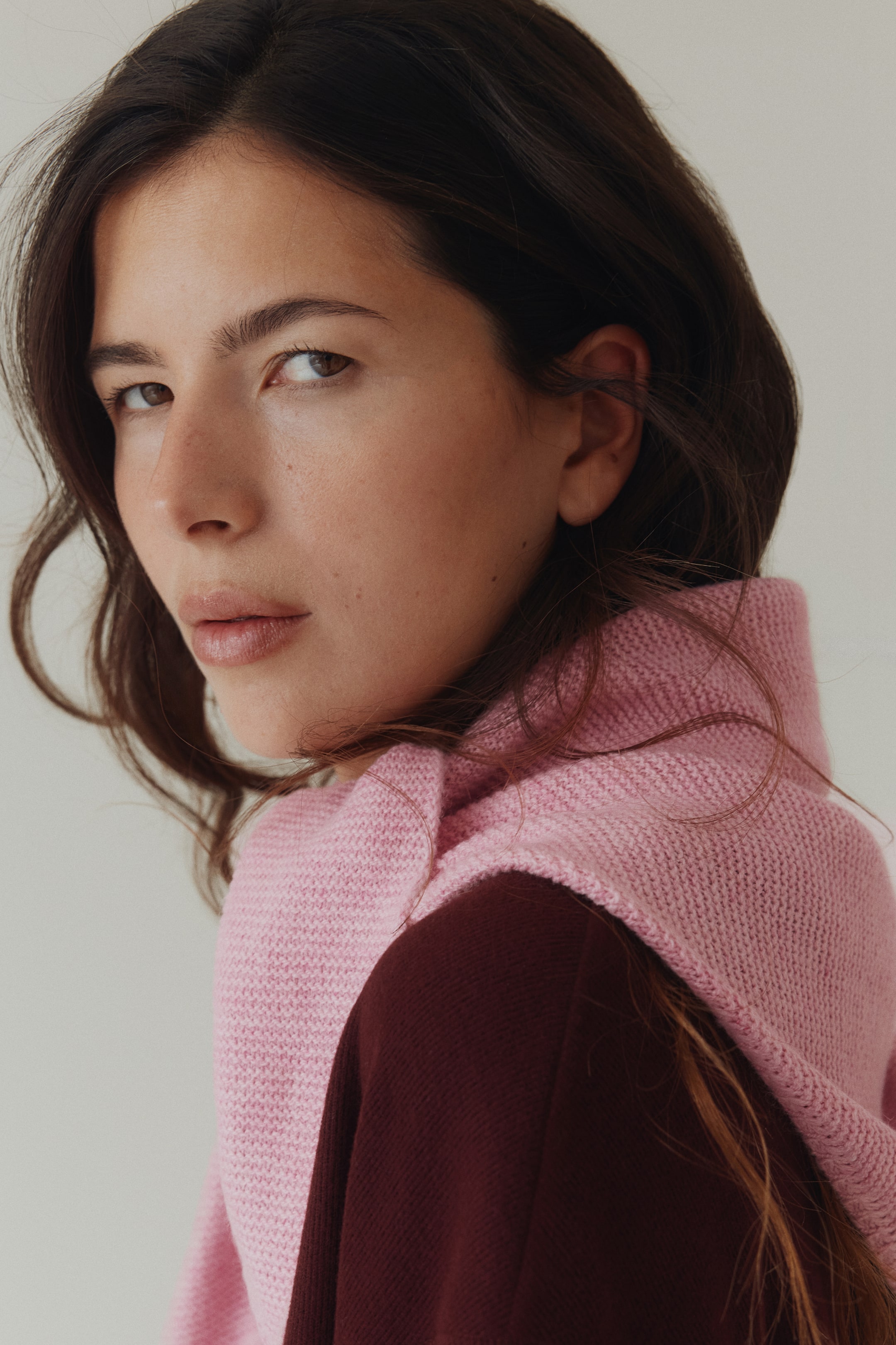 View larger image: A pink knitted cowl drapes over the shoulders and neck, showcasing a textured knit and layering over a dark red top. Shown from a slightly rear-left angle.