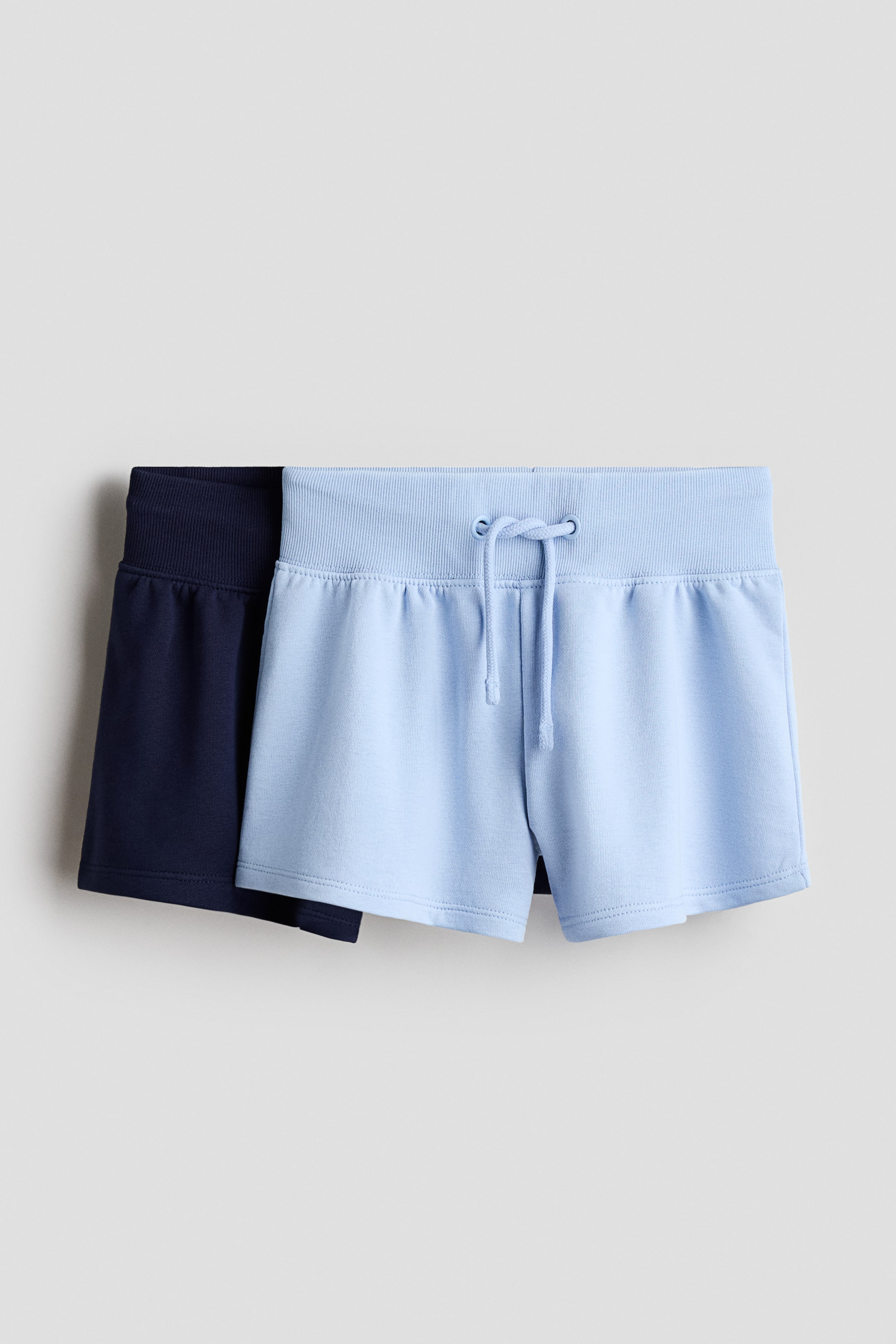 View larger image: Two pairs of loose-fit shorts made from lightweight sweatshirt fabric are displayed. The light blue pair, shown from the front, features a wide, ribbed waistband with a visible drawstring, while the navy blue pair is layered behind it.