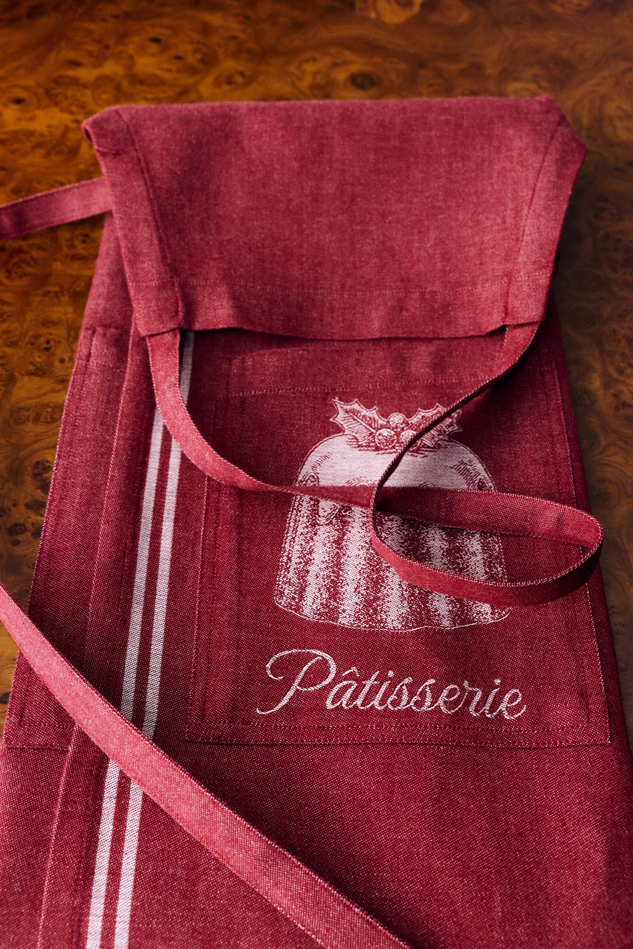 Se større billede: Et rødt forklæde i bomuldsvæv ligger fladt og viser en frontlomme med en hvid jacquard-illustration af en dessert toppet med kristtorn samt ordet "Pâtisserie" i kursiv skrift. Matchende smalle taljebånd ligger hen over forsiden, og to lodrette hvide striber løber ned langs siden.