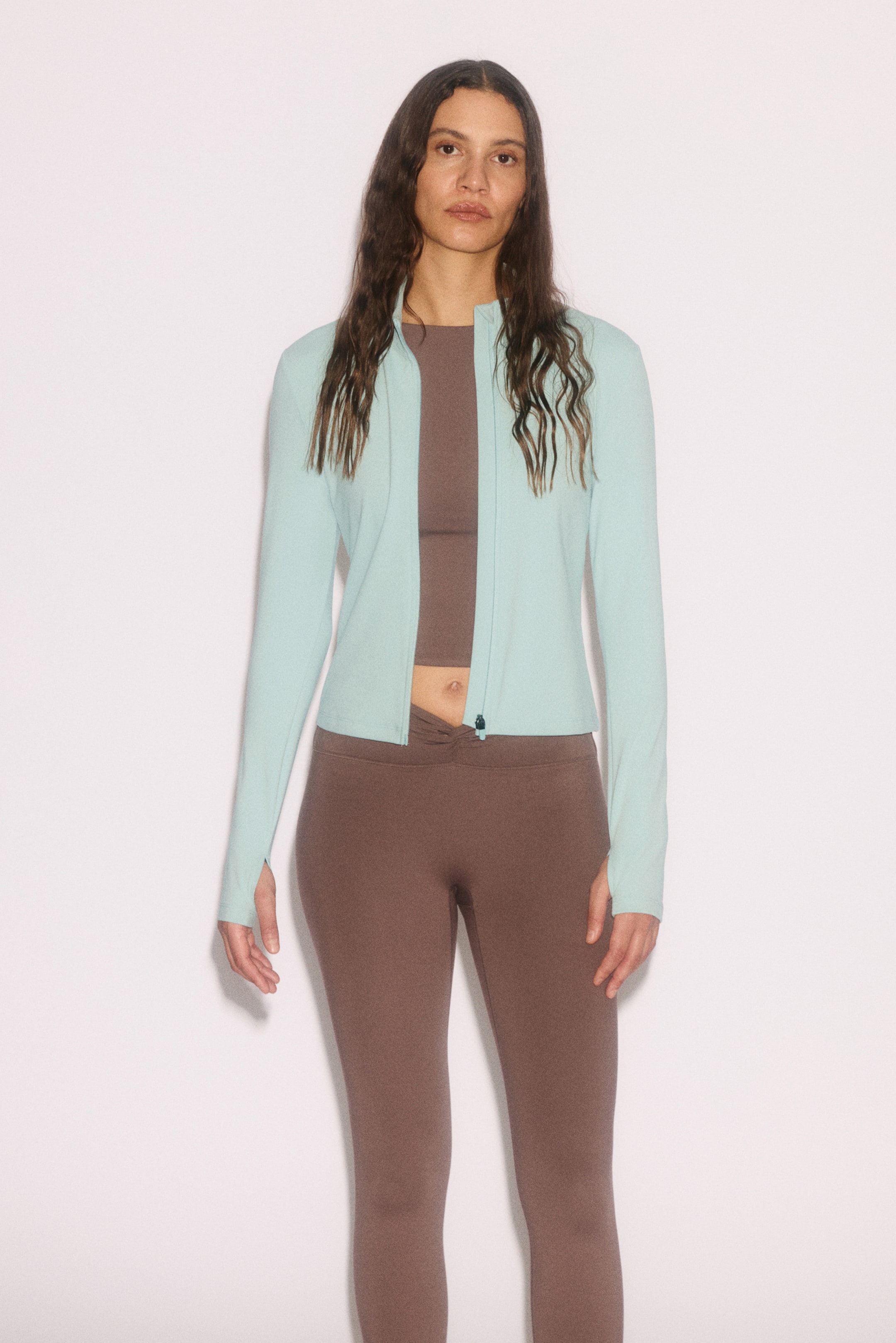 View larger image: "Sebuah jaket zip-up biru muda sedang dipersembahkan, menampilkan kolar berdiri dan lengan panjang dengan lubang ibu jari. Ia direka dengan zip depan penuh panjang dan dilapisi di atas crop top coklat, berakhir tepat di atas pinggang."