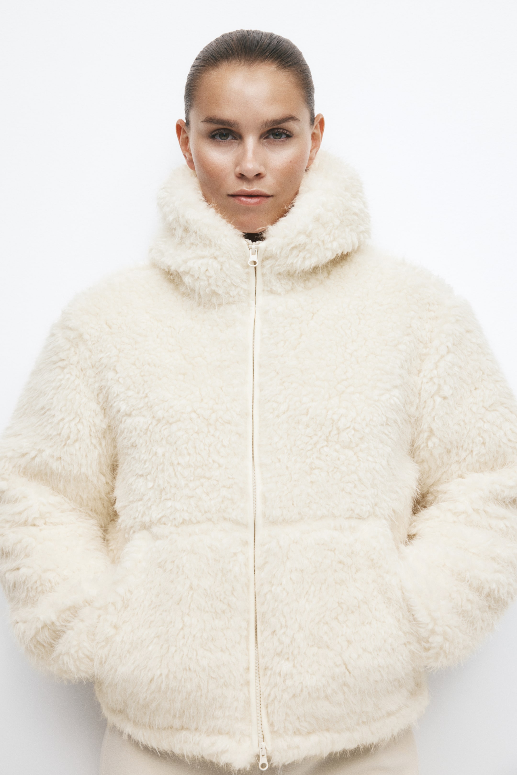 View larger image: A cream-colored, textured faux-shearling puffer jacket is worn by the model with their hands in the front pockets. The jacket features a high-collar hood and a full-length front zipper closure.