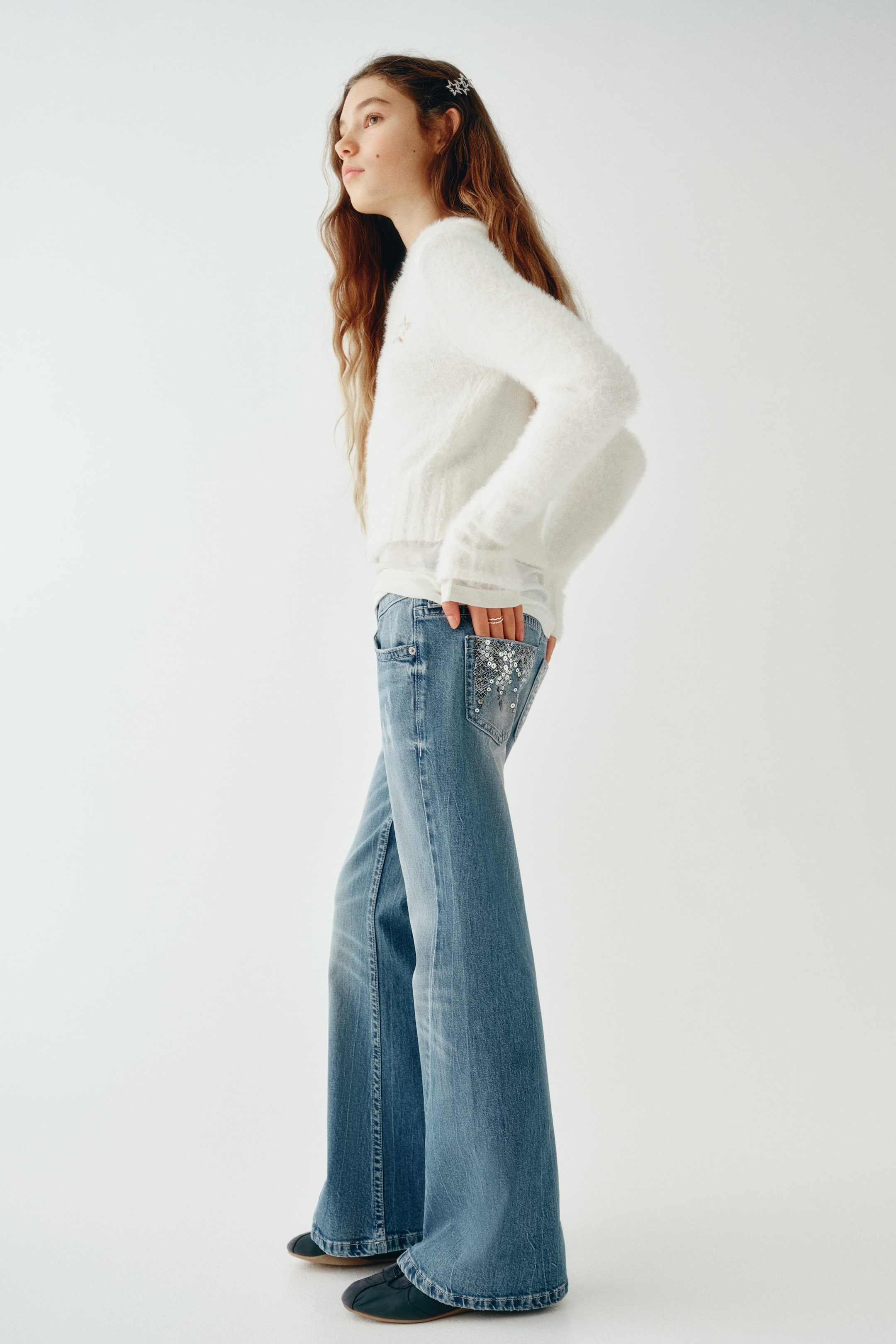 Büyük resmi görüntüle: Bu yıkanmış mavi pamuklu denim jeanler, rahat kesimlerini, kloş paçalarını ve burulmuş yan dikişlerini öne çıkaran yan profilden giyilmiş olarak görülüyor. Düşük belli jeanler, gümüş payetlerle detaylandırılmış işlemeli arka cep özelliğine sahip olup, beli vurgulayan yumuşak beyaz bir kazakla birlikte stilize edilmiştir.