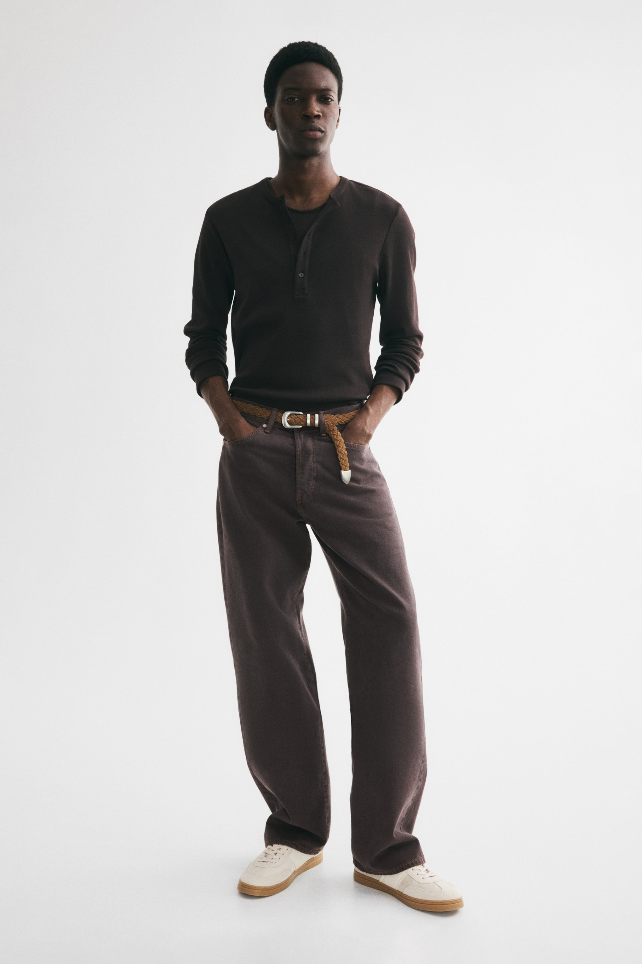 View larger image: The model is wearing dark brown 5-pocket jeans made from rigid cotton denim. They feature a regular waist, a dropped crotch, and a loose, rounded leg that offers extra room from the seat to the hem. Paired with these jeans is a braided belt, and the model's hands are casually placed in the front pockets.