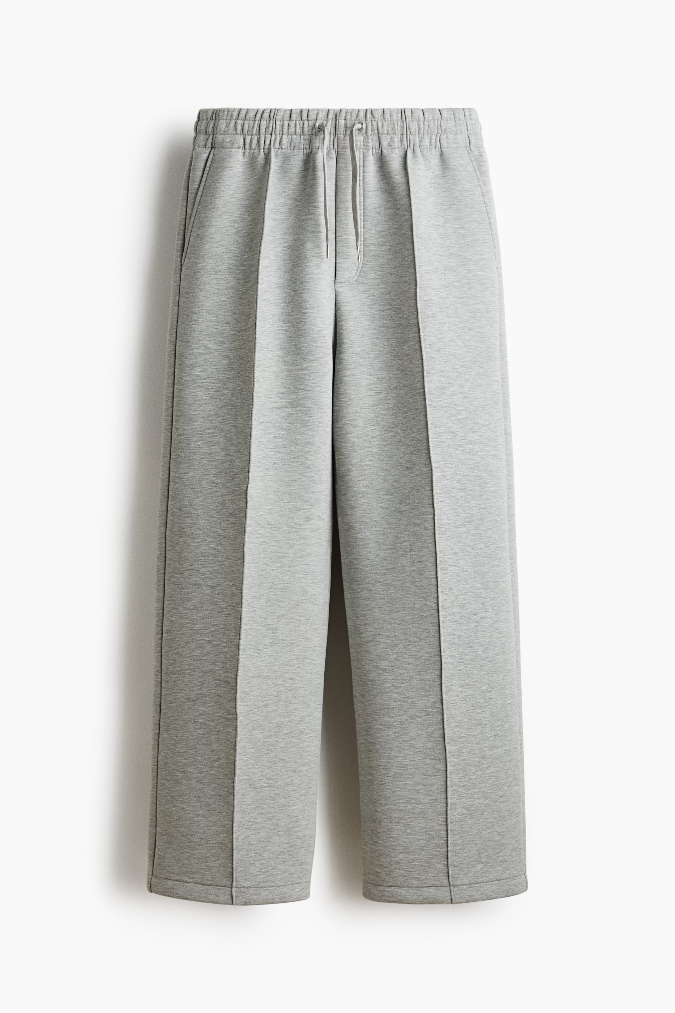 View larger image: Light grey scuba fabric joggers are displayed front-facing, highlighting an elasticated drawstring waist and a faux fly. Stitched creases run down the front of each leg, with side pockets visible, offering a relaxed, straight-leg silhouette.