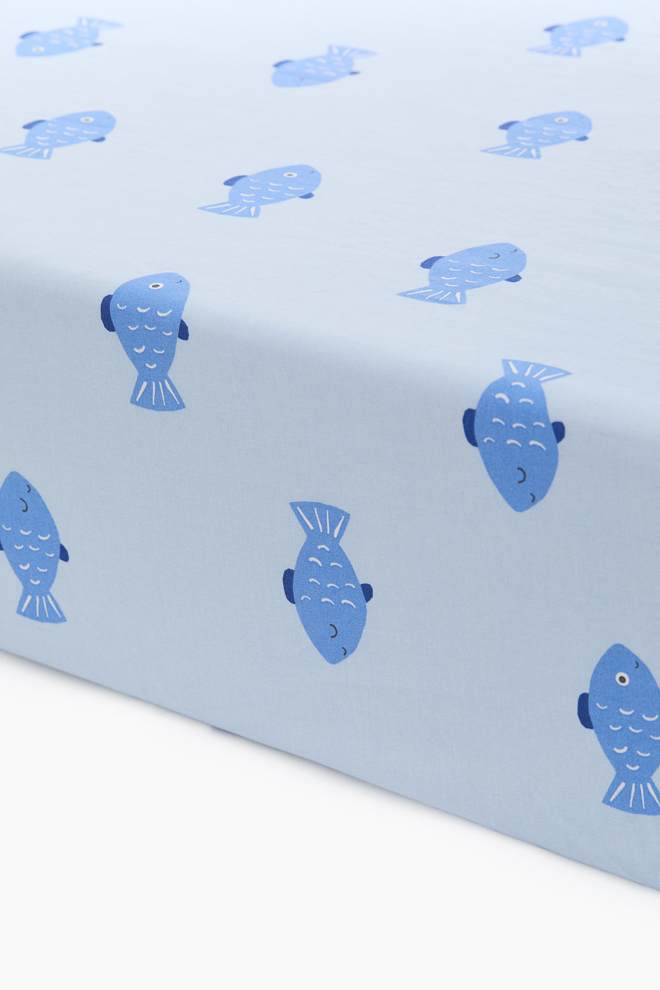View larger image: A fitted sheet in a light blue patterned cotton weave is showcasing its top and side surfaces. The sheet features a repeating pattern of dark blue cartoon fish swimming in various directions.