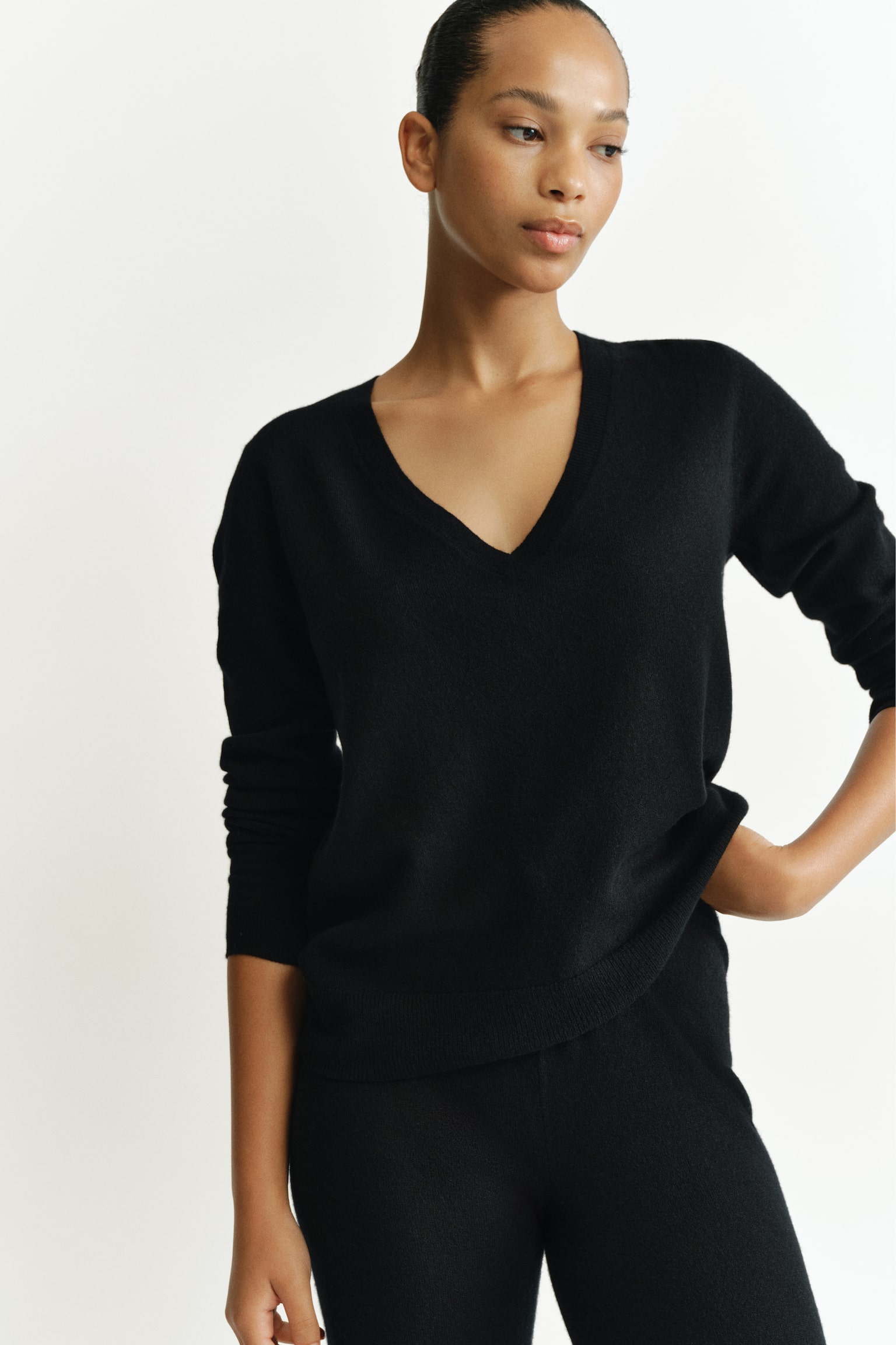 A black wool-knit jumper featuring a deep, rib-trimmed V-neck and dropped shoulders is worn by the model. The model is pairing the long-sleeved jumper with matching black knit trousers, standing with one hand resting on the hip. - 1