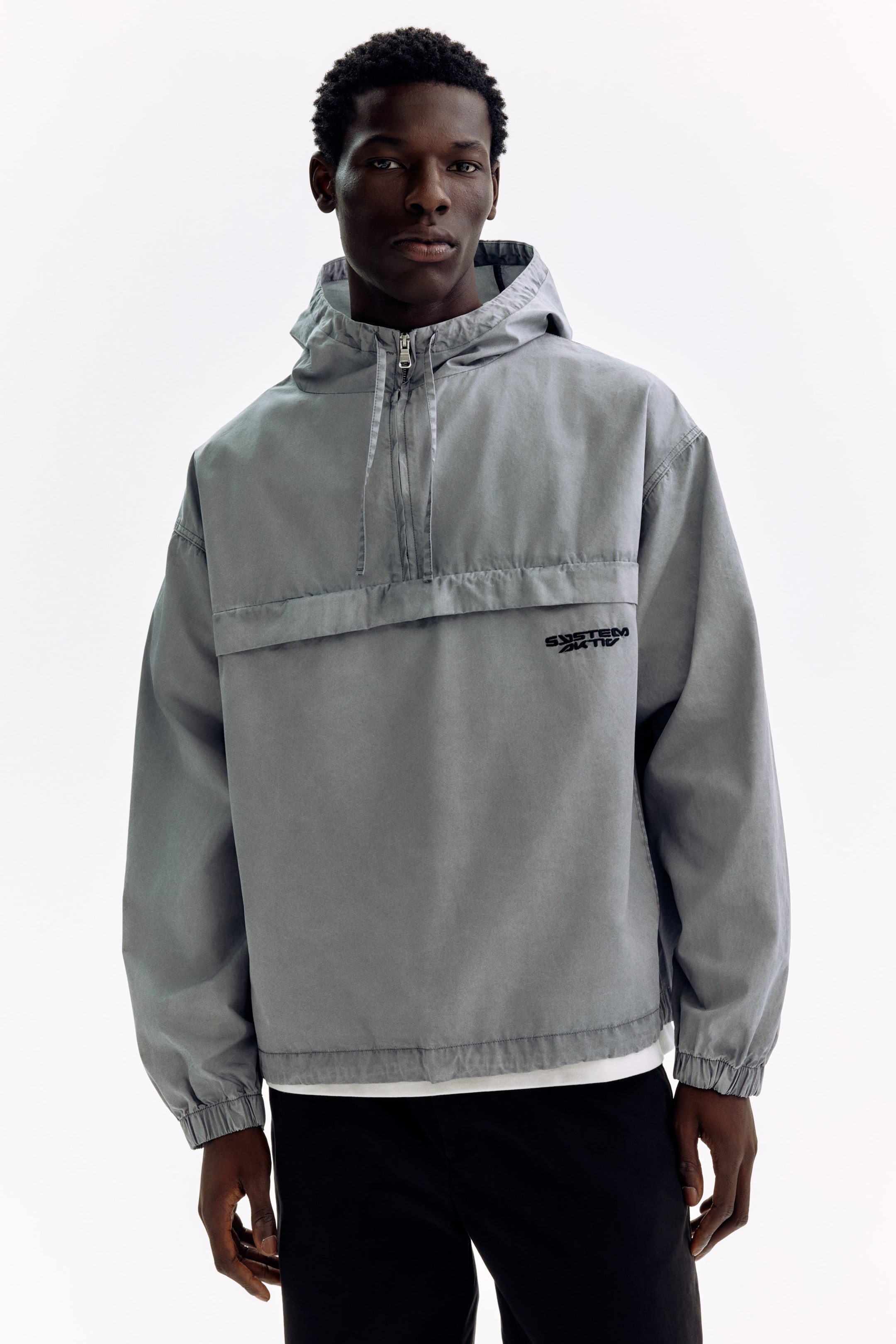 Ver imagen más grande: La chaqueta anorak gris está de frente, con capucha y cierre de media cremallera. Presenta puños y dobladillo elásticos, un panel horizontal en el pecho y el texto negro 'SYSTEM AKTIK' en el lado izquierdo del pecho del usuario. Se ve una camiseta blanca debajo del dobladillo de la chaqueta.