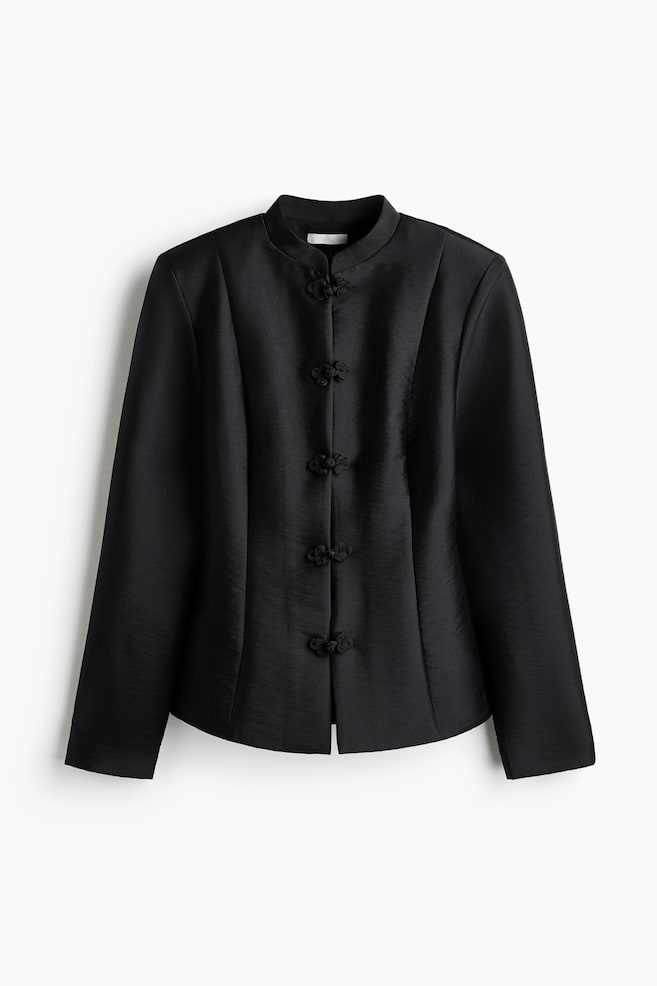 A black structured jacket hangs facing forward, featuring a band collar and five decorative knot-and-loop closures down the centre. Vertical seams run down the front of the subtly lustrous fabric, creating a tailored shape with long sleeves. - 1