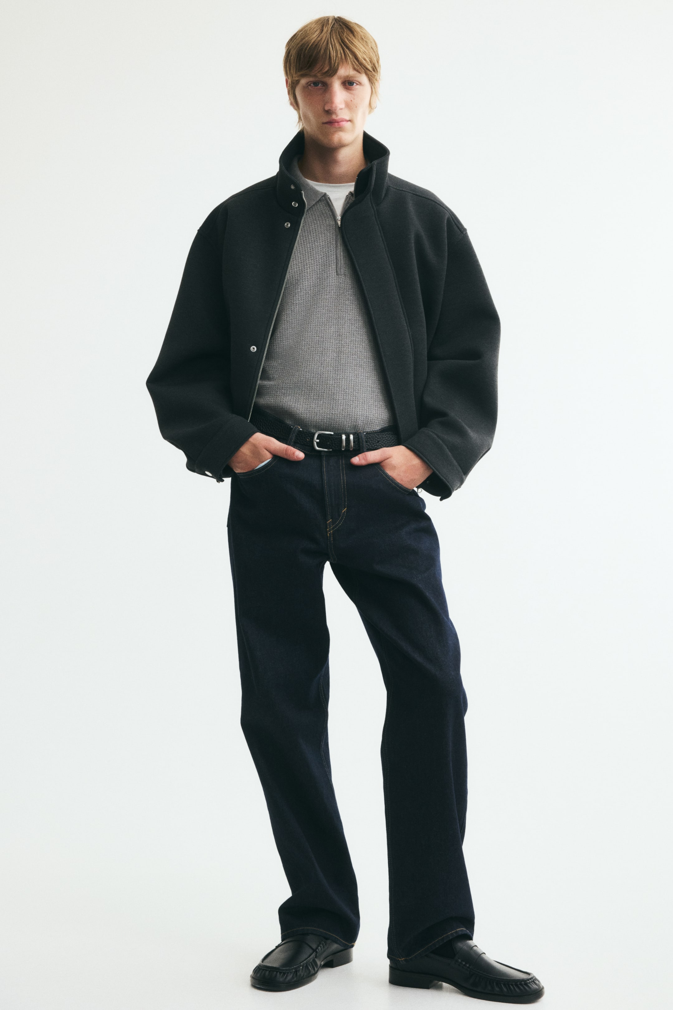 View larger image: A light grey textured knit polo shirt, featuring a ribbed collar and a partially unzipped zip at the top, is being worn by the model. The model is layering the shirt under a dark jacket.