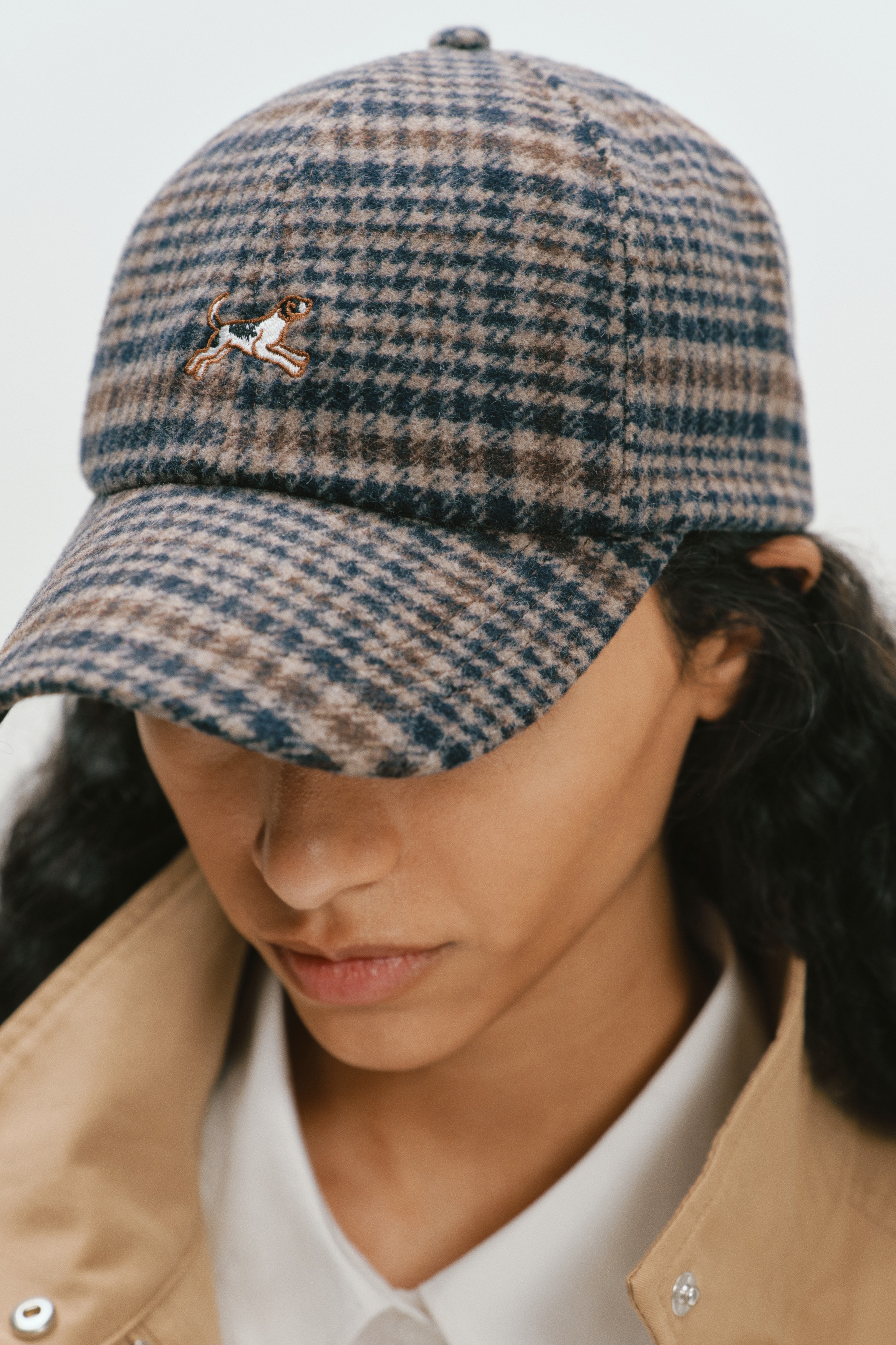 Vista frontal de una gorra de bÃ©isbol de mezcla de lana con un estampado de cuadros en marrÃ³n, negro y gris, y un acabado suave y texturizado. La gorra muestra un pequeÃ±o perro corriendo bordado en el centro frontal de la copa.
