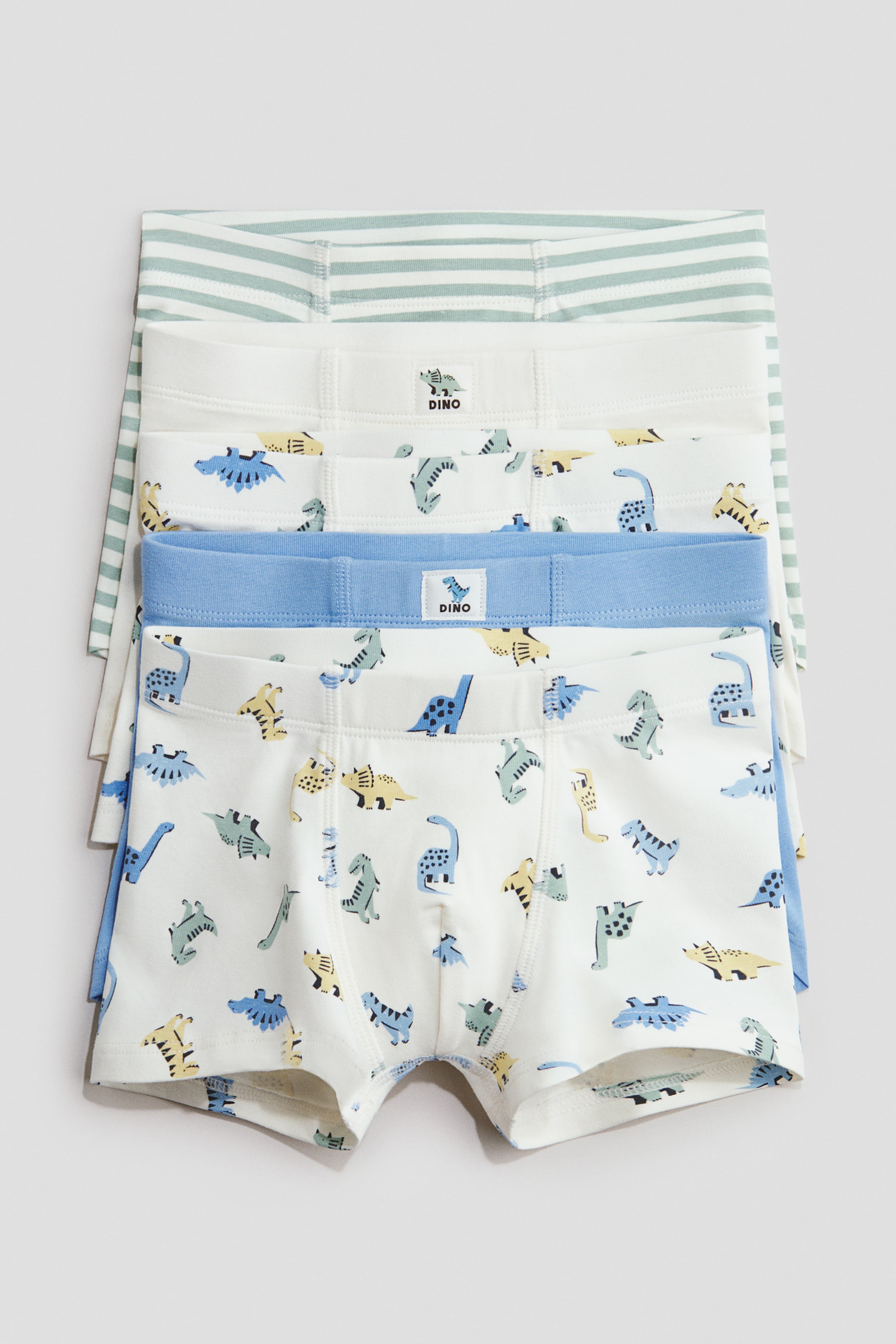Agrandir l'image: Quatre paires de shorts en jersey de coton sont empilées, présentant leur devant avec des jambes courtes, des ceintures élastiquées, et des devants doublés. La paire du haut présente des rayures horizontales vert clair et blanches, tandis que les trois autres paires présentent deux paires blanches avec des motifs de dinosaures de dessins animés bleu, jaune et vert, accompagnées d'une paire unie bleu clair.