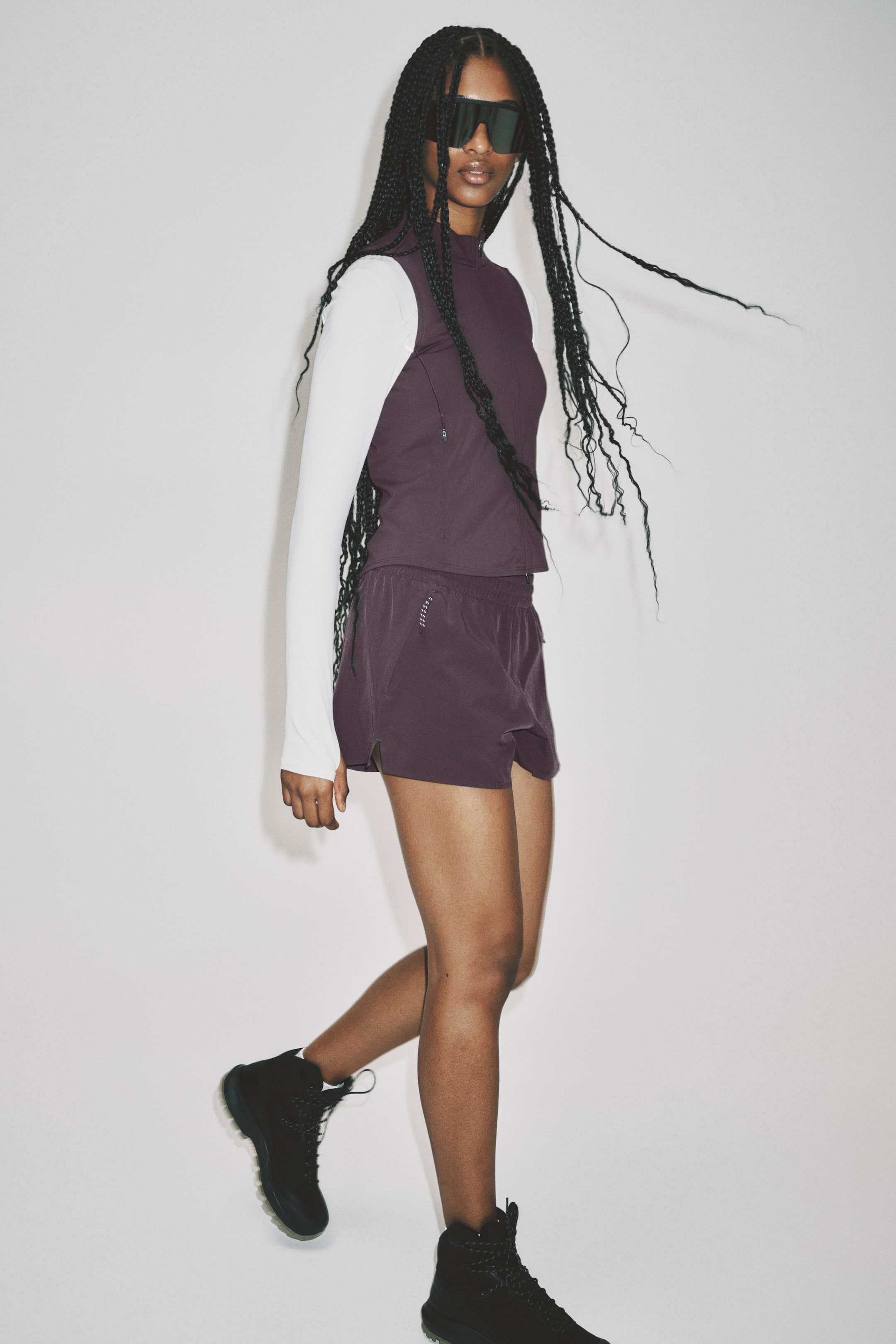 View larger image: The ensemble highlights a dark plum sleeveless mock-neck top featuring a visible chest zipper, elegantly layered over a long-sleeved white base layer. Coordinating dark plum athletic shorts complete the lower half, accented by a subtle reflective detail on the left thigh. Black high-top sneakers with rugged soles finish the look.