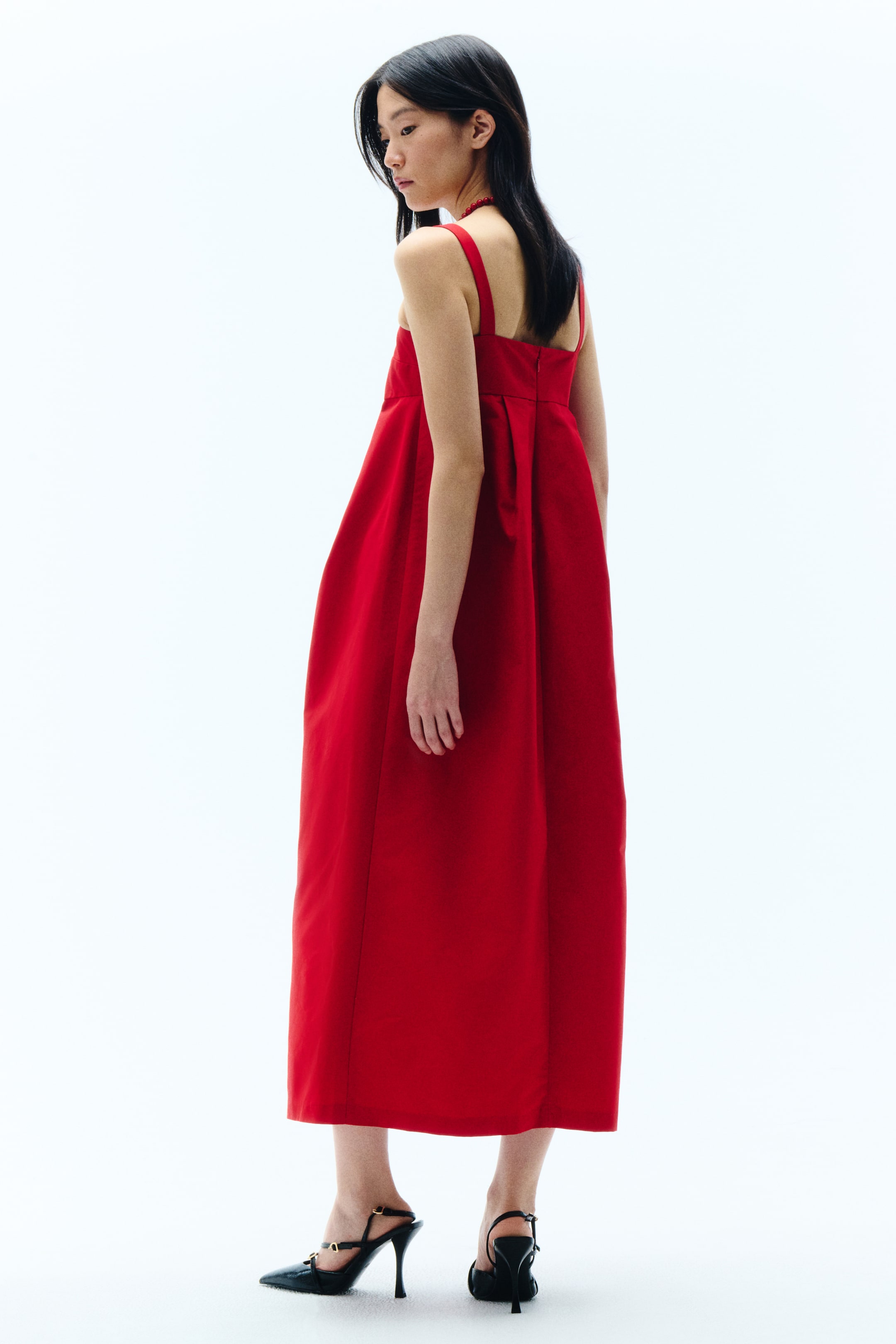 View larger image: A red midi dress with wide shoulder straps and a voluminous skirt featuring visible pleats at the back, shown from a back three-quarter angle.