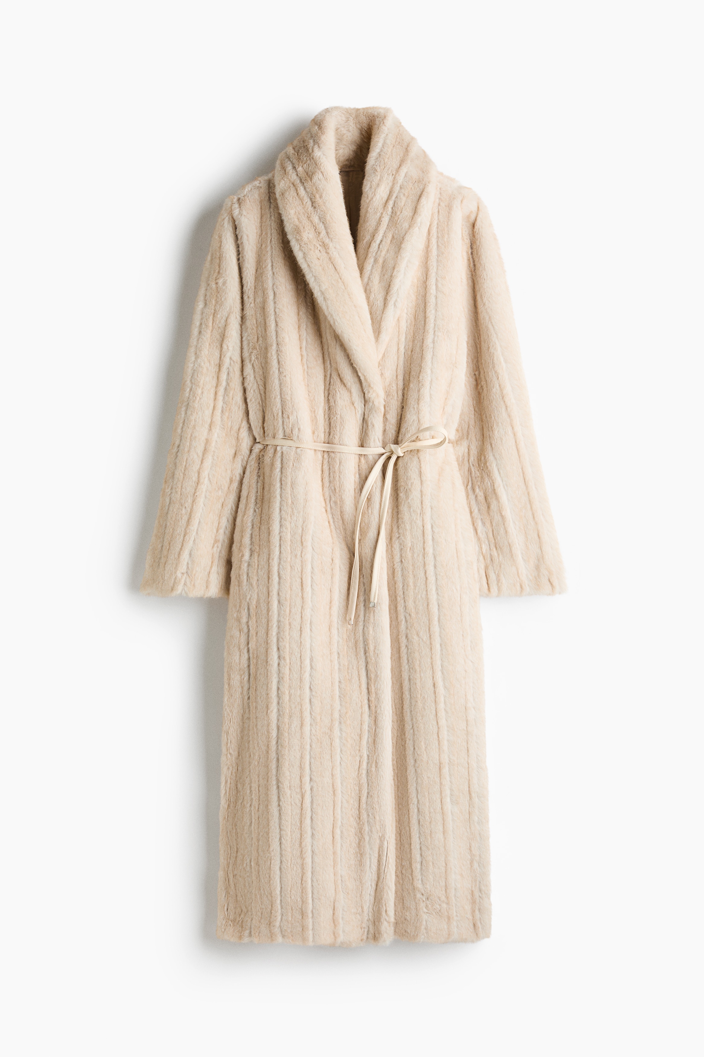 A cream-colored, fluffy maxi coat featuring a textured finish, a shawl collar, and a thin tie belt at the waist is being worn by the model. The model is posing with hands placed in the side pockets, layering the coat over a white top, blue jeans, and sneakers.