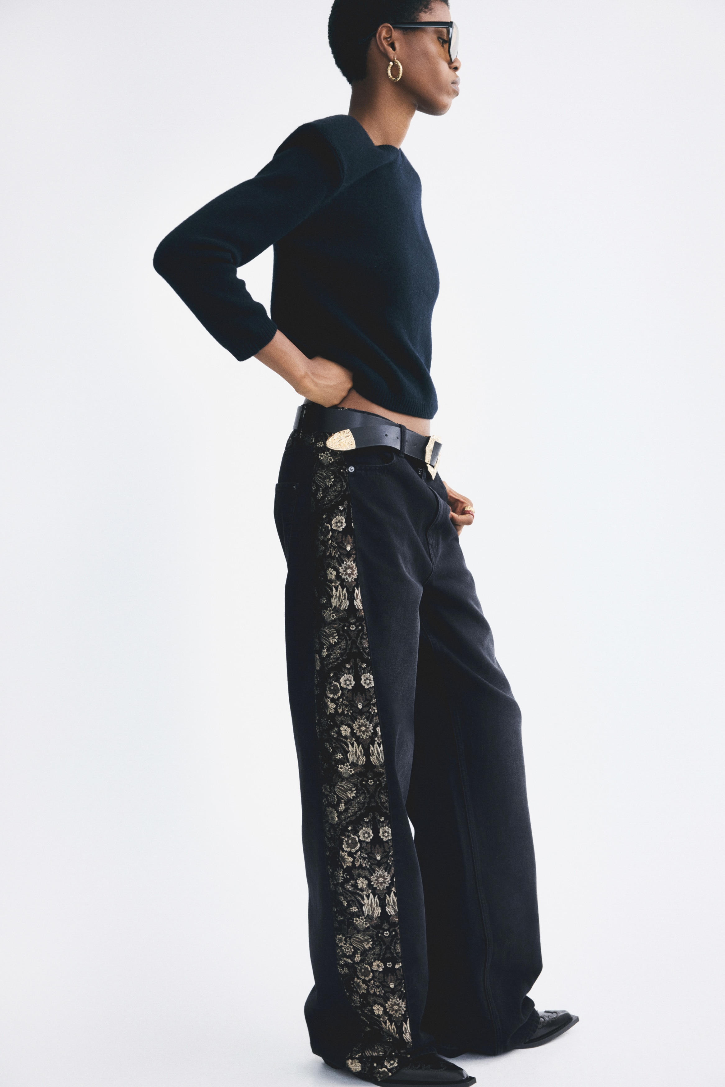 A pair of wide-leg black denim jeans is facing front, showcasing contrasting metallic-gold floral brocade panels running down both sides. The design features a classic button-and-zip fly fastening, belt loops, and curved front pockets.