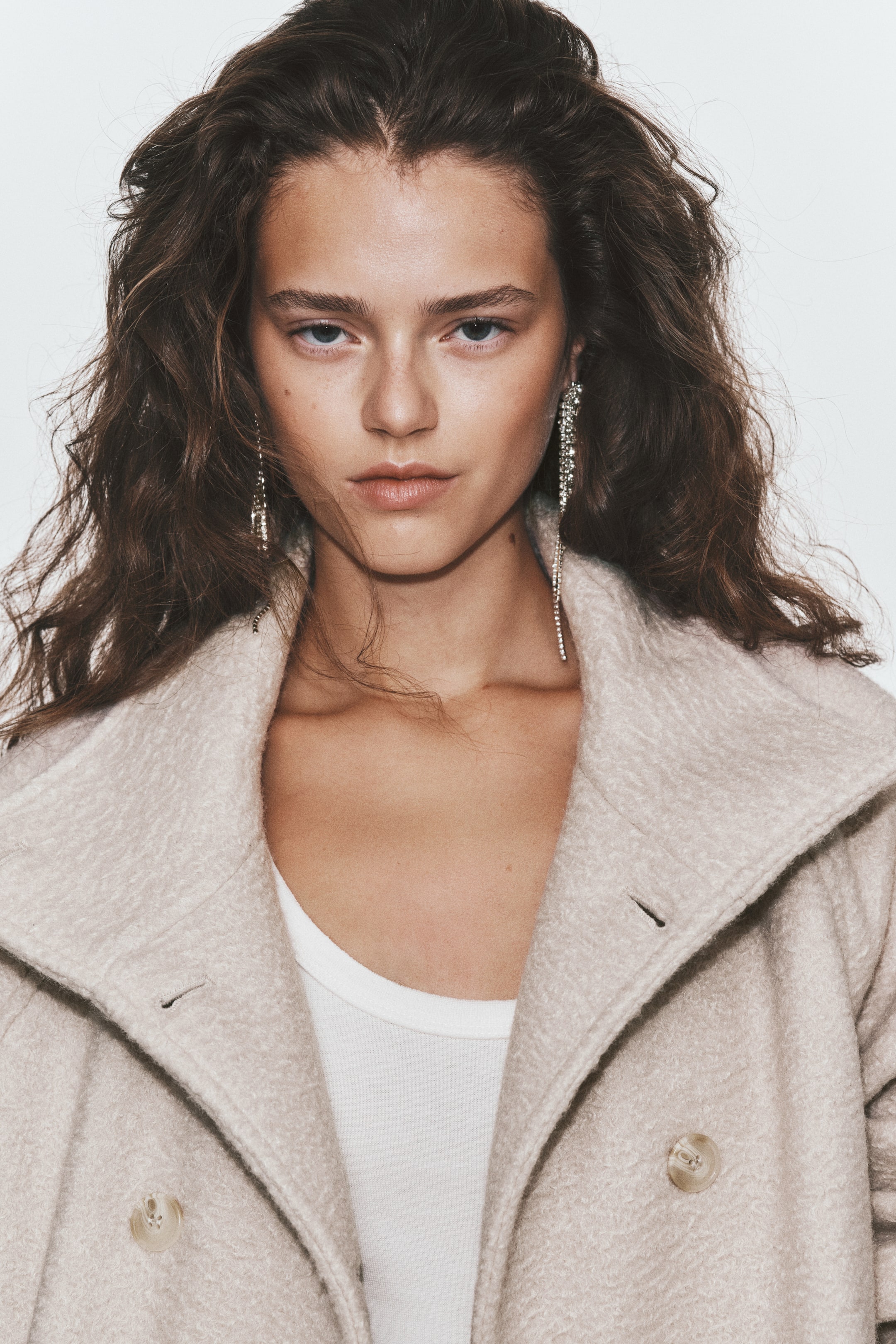 View larger image: A double-breasted jacket in a light beige felted fabric is worn by the model over a white top. The jacket features a wide collar worn open, with light-coloured front buttons and buttonholes visible.
