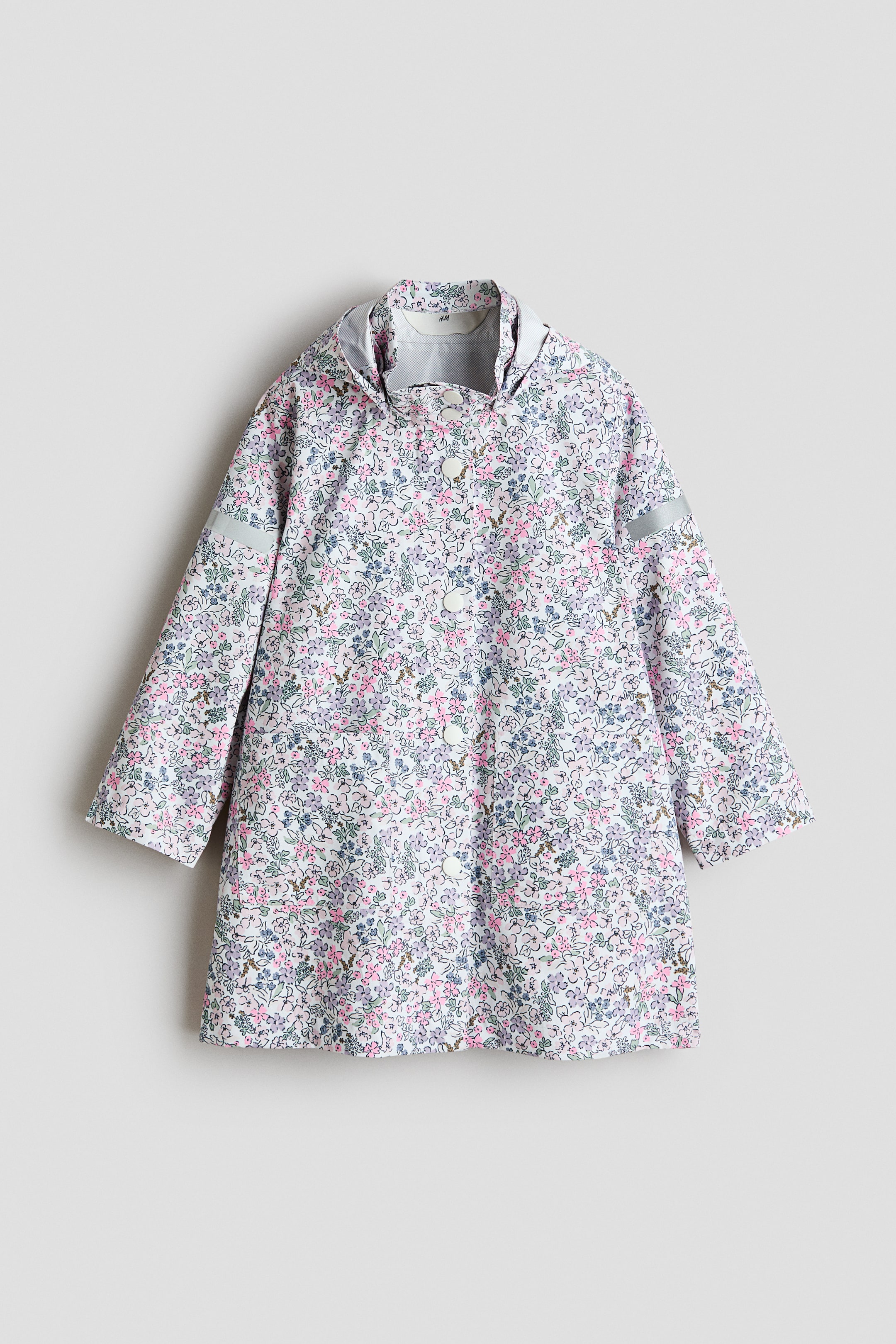 Grotere afbeelding bekijken: Een knielange regenjas presenteert zijn voorkant, met een witte basis en een algehele bloemenprint in roze, paars en groen. Hij is ontworpen met een opstaande kraag, een afneembare capuchon, een voorflap tegen de wind die vastgezet wordt met witte drukknopen, twee voorzakken en reflecterende strips op beide mouwen.