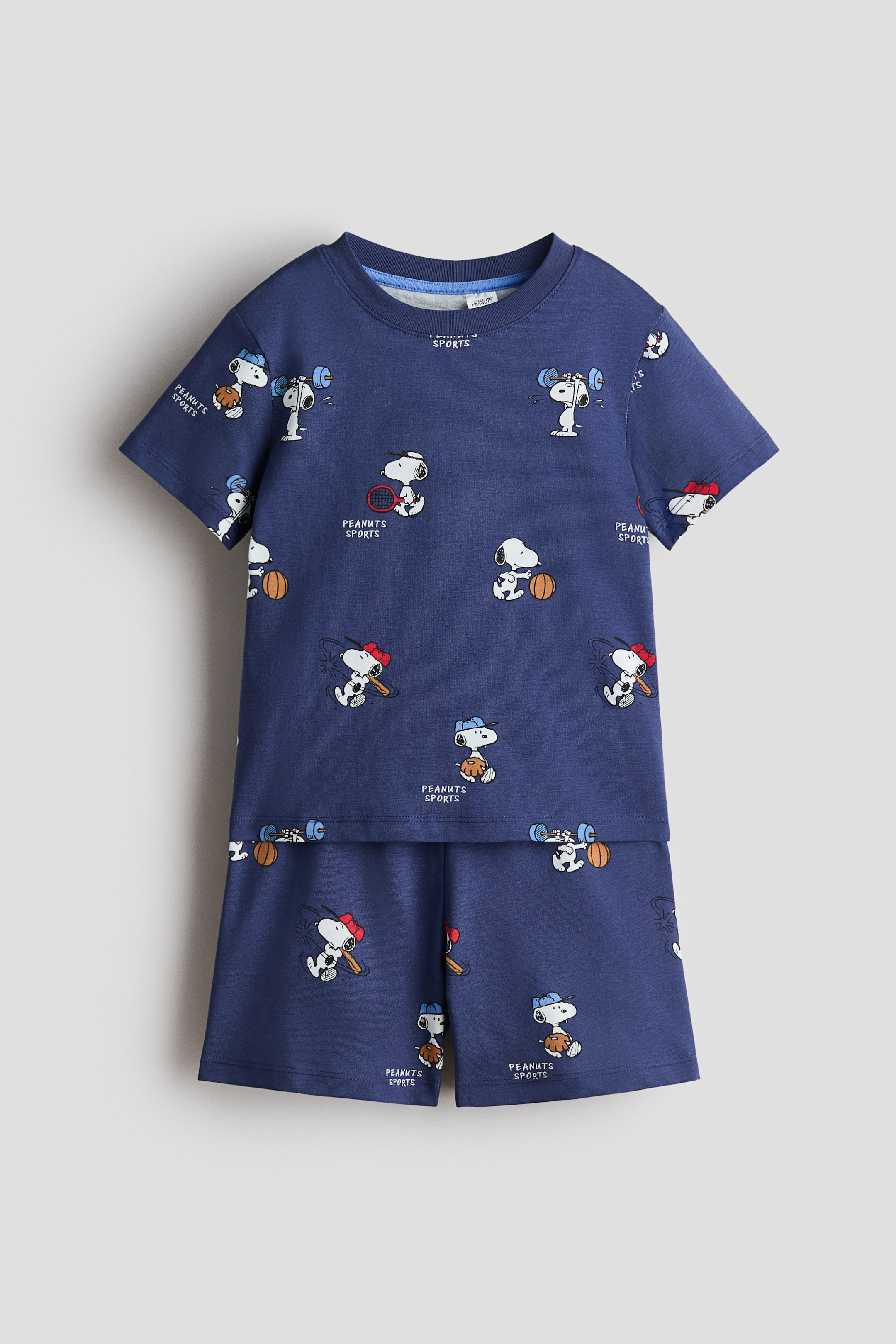 Ver imagem maior: Um conjunto de pijama azul-marinho está a exibir a sua parte frontal, com uma t-shirt com um estampado desportivo do Snoopy por toda a peça, mangas curtas e um acabamento canelado em torno da gola. Os calções a condizer apresentam o mesmo estampado e uma cintura elástica.