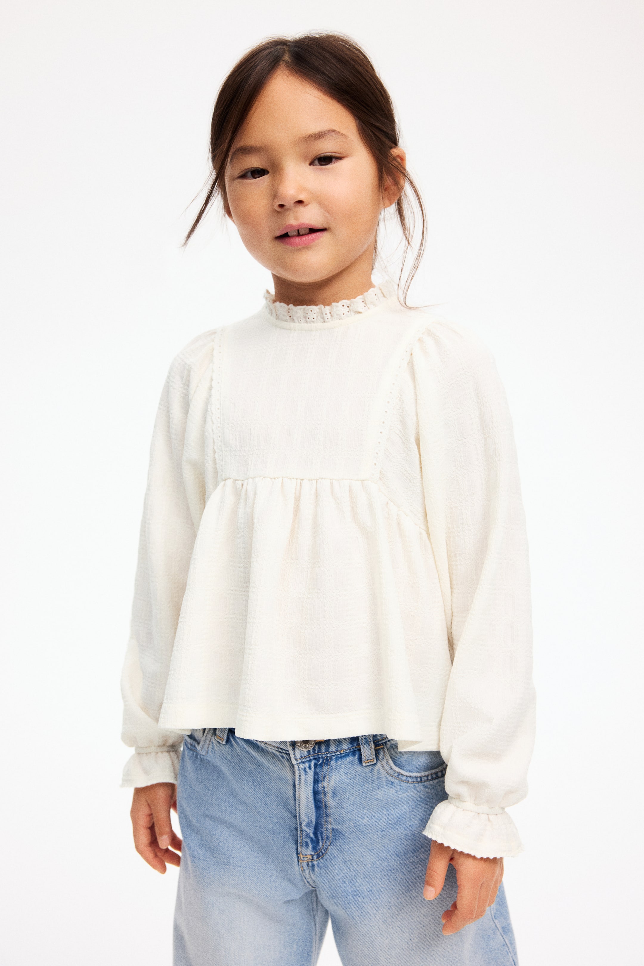 拡大画像を表示: ホワイトの長袖トップスは、表面にテクスチャーのある生地とハイネックのフリル襟があしらわれています。パフショルダー、身頃の縦ラインに入ったレーストリム、ギャザーを寄せたペプラムヘム、フリル付きのゴム入りカフスが特徴です。
