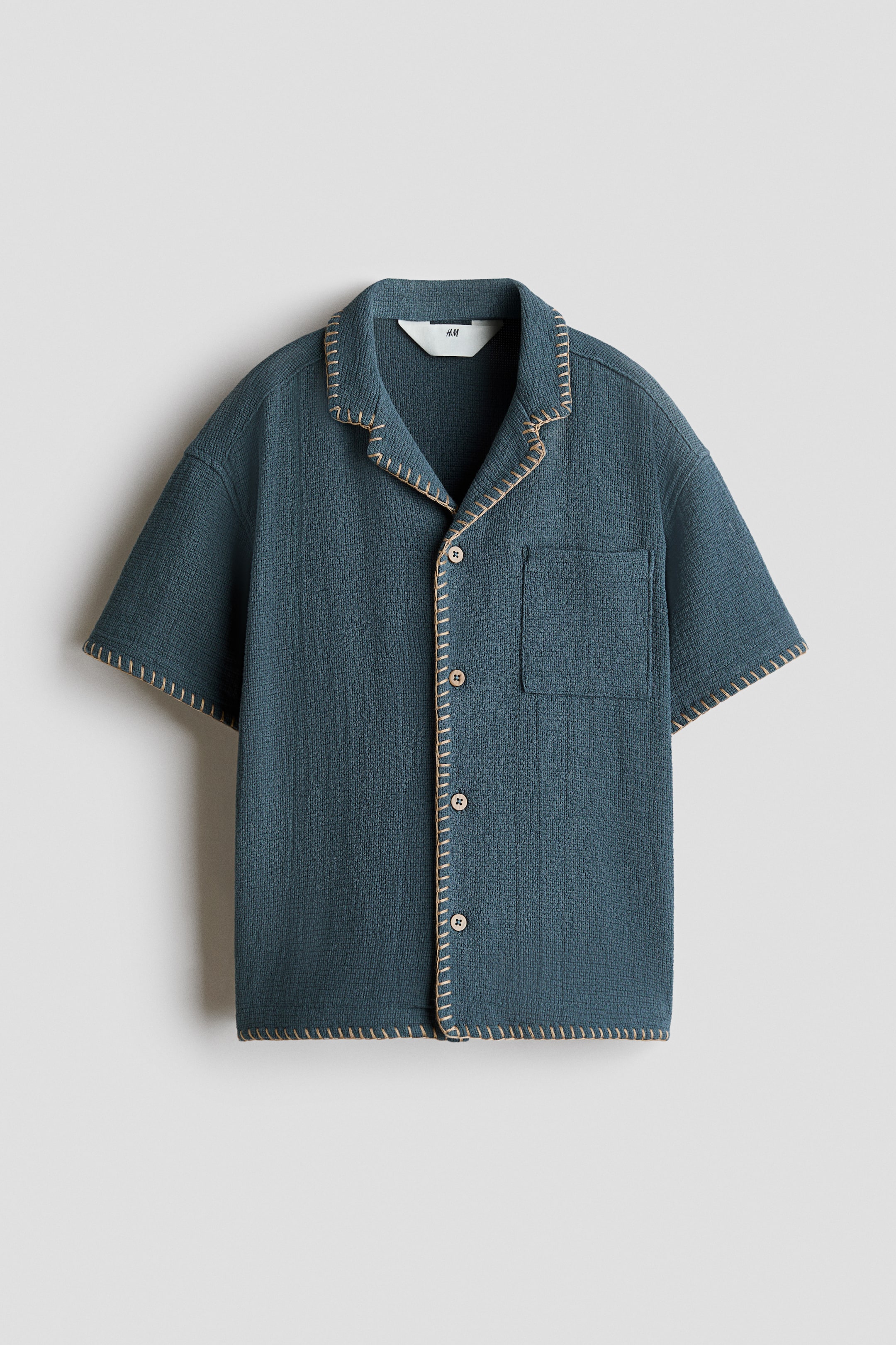 View larger image: A dark teal short-sleeved shirt is presented front-facing, featuring a textured waffle-knit fabric and a camp collar. It is designed with a button-up front with light-coloured buttons, a single patch pocket on the left chest, and contrasting beige blanket stitching along all edges.