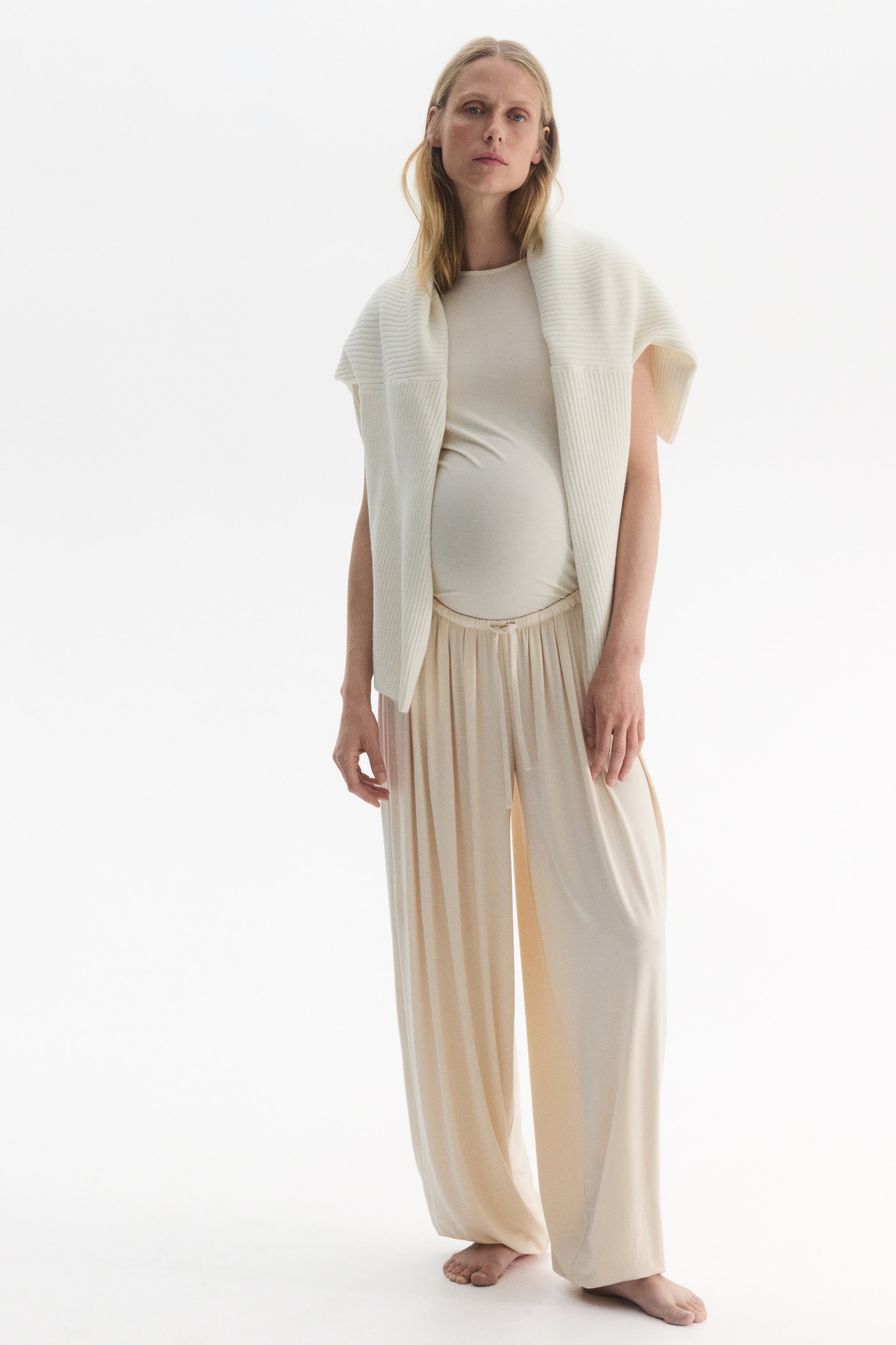 Visualizza l'immagine ingrandita: Un completo in jersey di viscosa elasticizzata color crema, indossato dalla modella, composto da una t-shirt a girocollo aderente e pantaloni morbidi e fluidi con vita elasticizzata con coulisse. Un maglione coordinato a maglia a costine è appoggiato sulle spalle della modella come capo a strati.