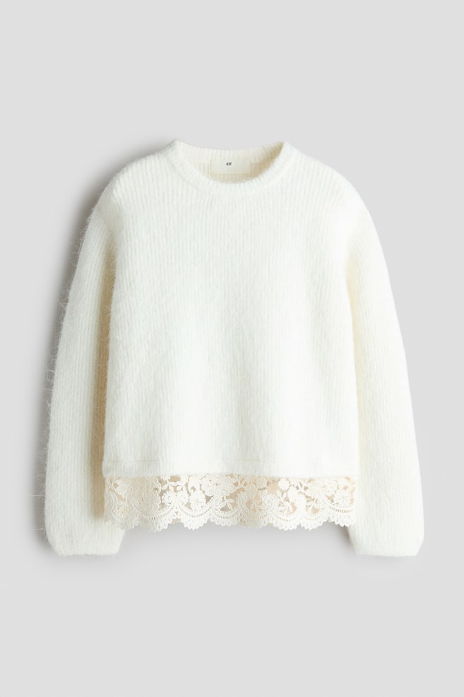 A cream-coloured knit jumper, facing front, features a soft, fuzzy texture with a ribbed crew neck and long sleeves. The garment is displaying a delicate, scalloped floral lace trim along its bottom hem. - 1
