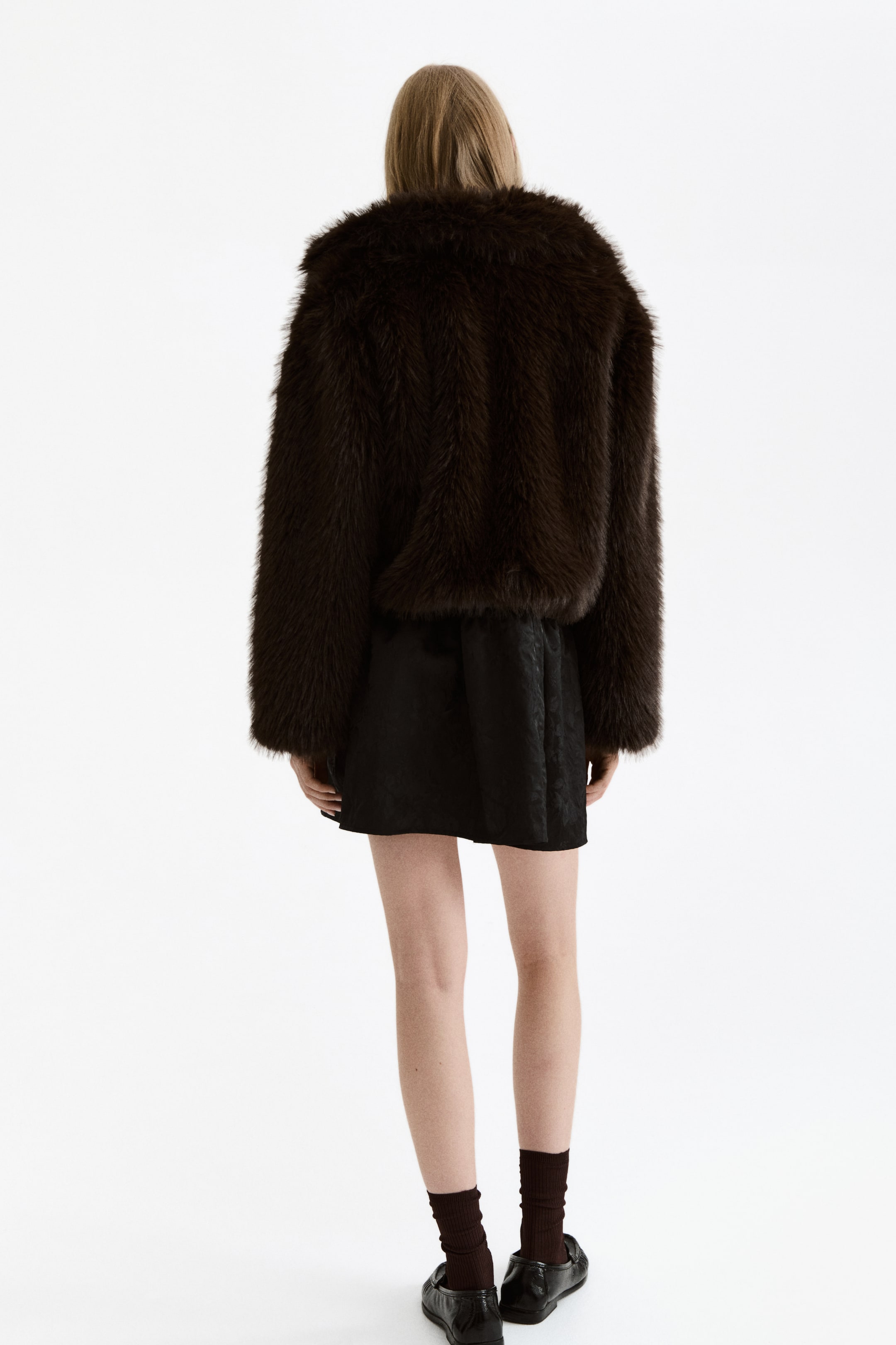View larger image: A brown faux fur jacket is being presented from the back view, featuring a shaggy texture and long, full sleeves. The cropped jacket ends at the upper hip, layered over a black skirt.