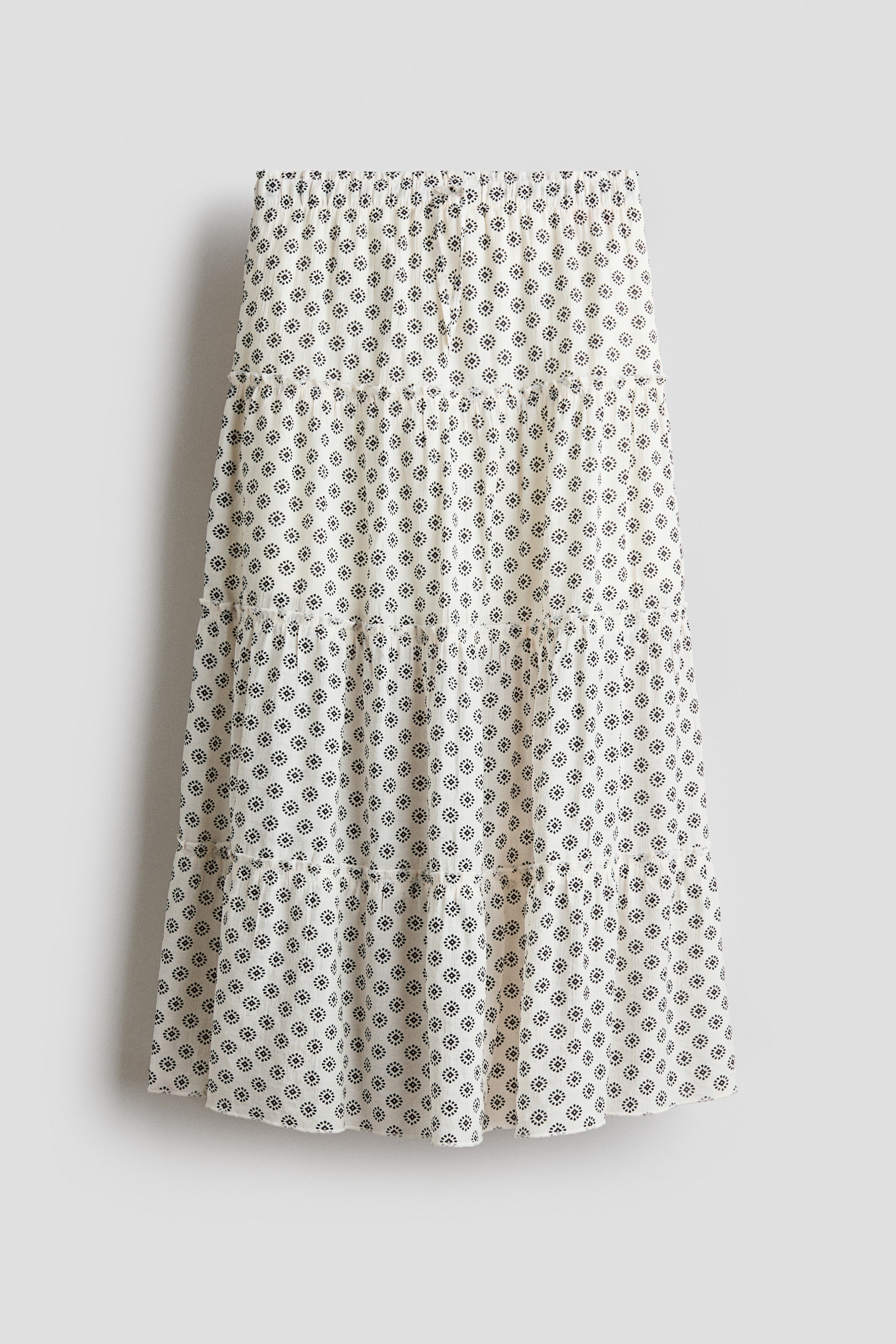 View larger image: The front of an off-white tiered skirt showcases a repeating black floral pattern. It features a gathered elastic waistband and multiple gathered tiers.