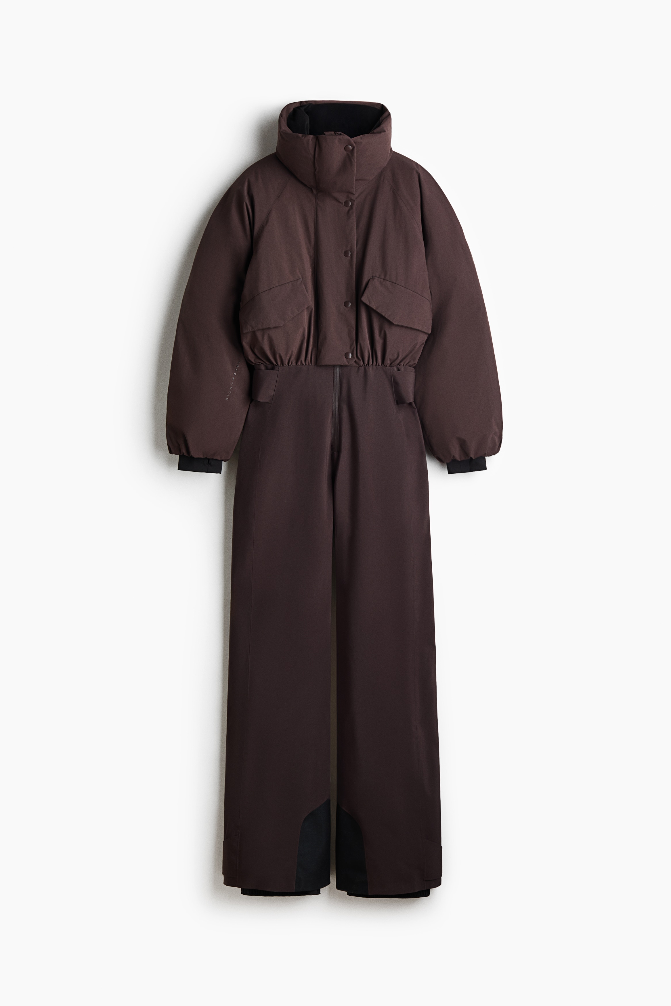 Un ensemble de ski marron foncÃ©, composÃ© dâune doudoune courte avec un col montant Ã  effet entonnoir et dâun pantalon taille haute assorti, est portÃ© par le mannequin. La veste prÃ©sente une fermeture pressionnÃ©e sur le devant et des poches Ã  rabat, et est associÃ©e Ã  des bottes de ski noires.