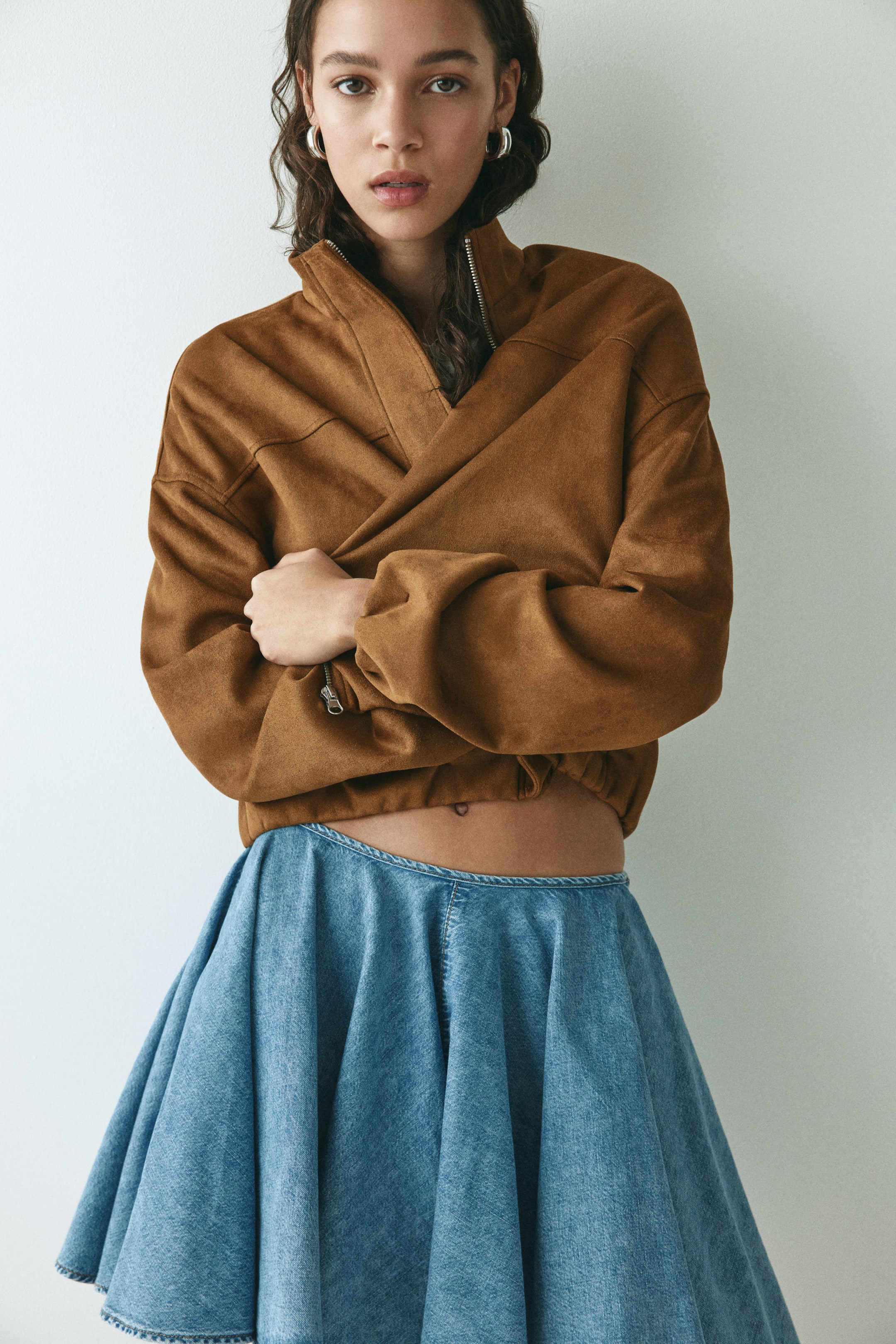 Προβολή μεγαλύτερης εικόνας: Ένα καφέ cropped μπουφάν φαίνεται από μπροστά, αναδεικνύοντας ψηλό γιακά, ορατό ασημένιο φερμουάρ και μακριά, χαλαρά μανίκια με ελαστικό τελείωμα. Το συμπληρώνει μια denim φούστα σε ανοιχτό πλύσιμο, που κάθεται ψηλά στη μέση και ανοίγει προς τα κάτω.