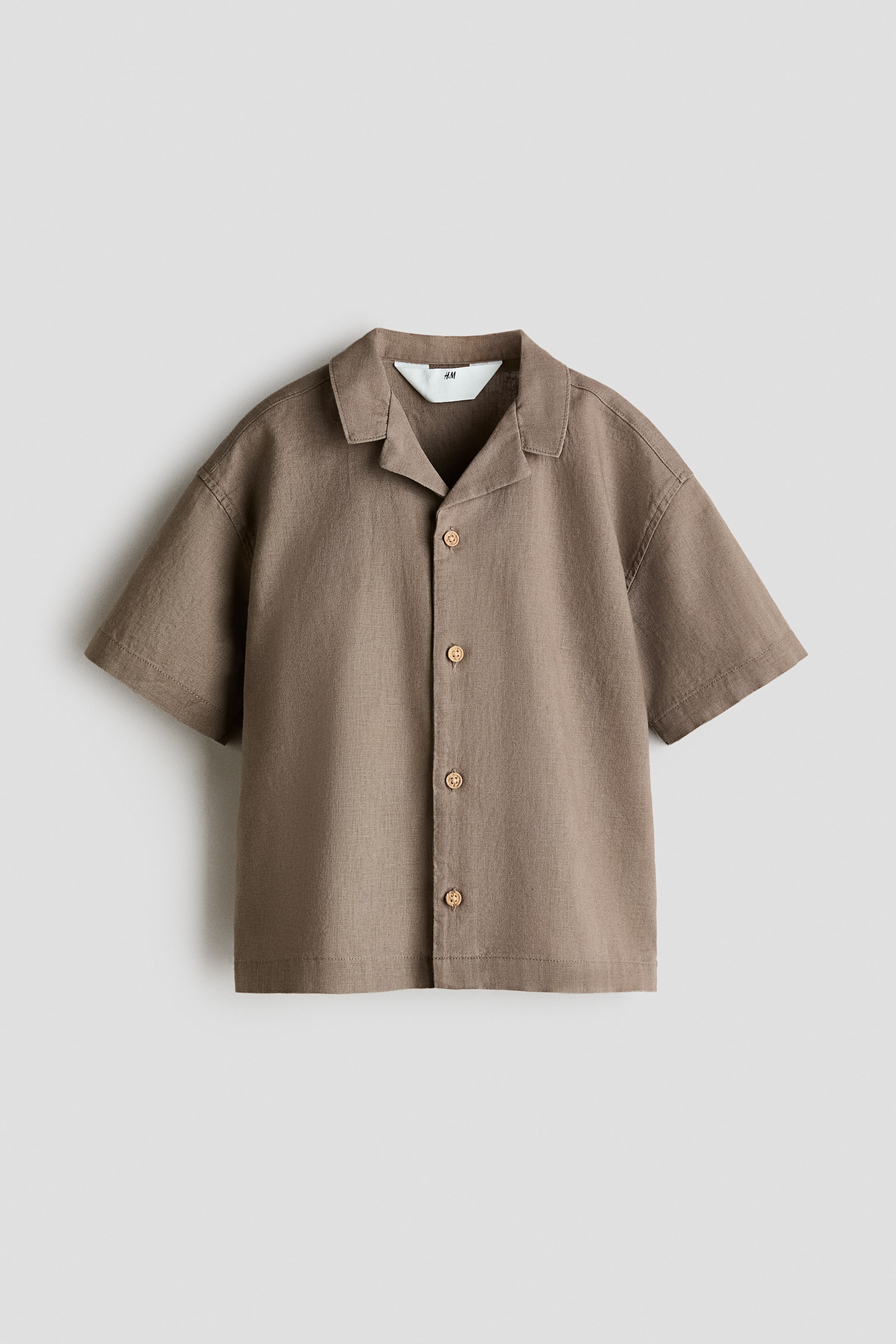 Ver imagem maior: "Uma camisa de mangas curtas em uma mistura leve de linho e algodão exibe a sua frente, com uma gola resort, ombros ligeiramente caídos e quatro botões redondos ao longo da frente."