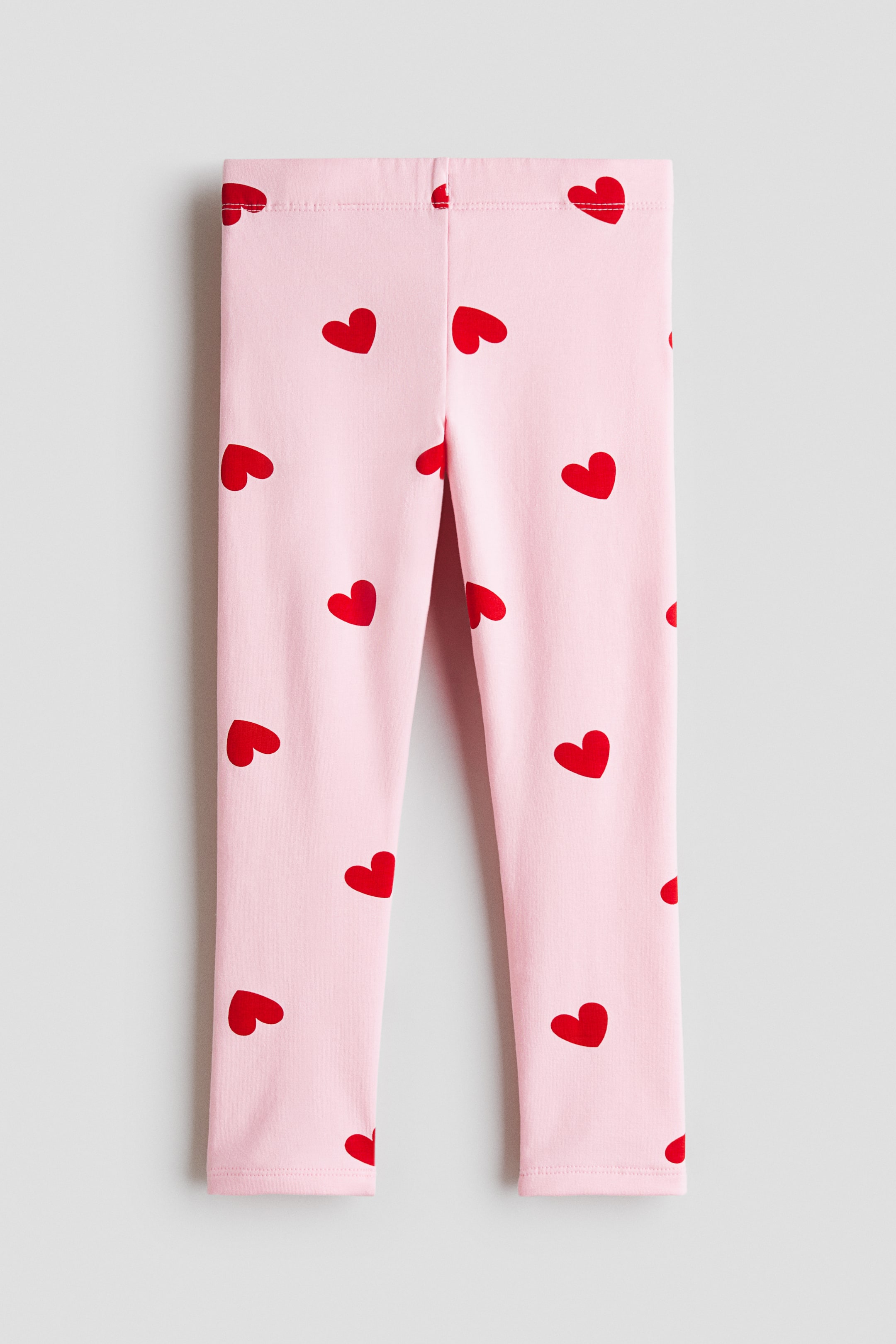 View larger image: The leggings feature a soft pink hue adorned with a repeating motif of red hearts. Displayed from a front perspective, they boast an elasticated waistband.