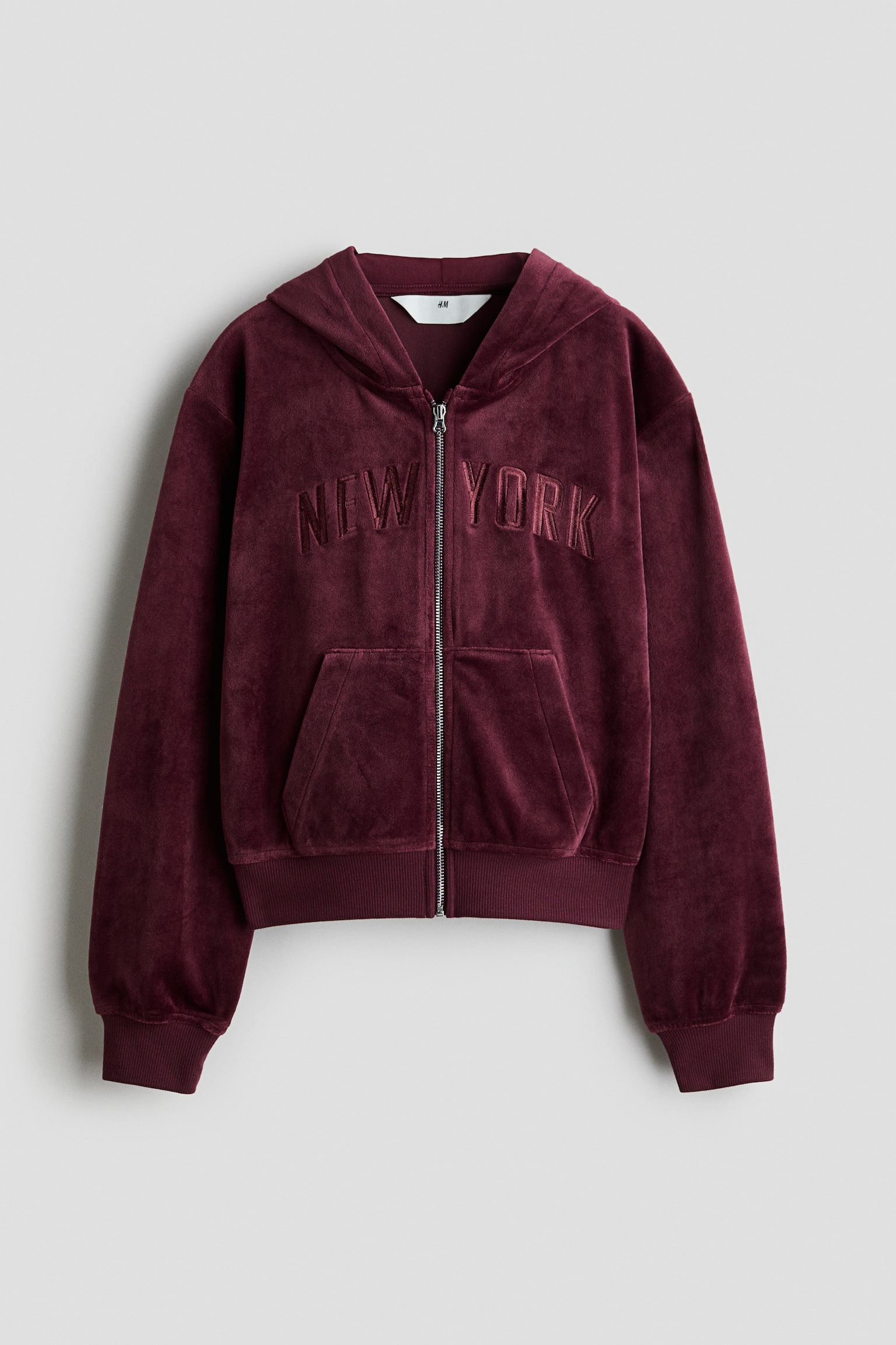 A burgundy velour zip-up hoodie is facing front, featuring the text "NEW YORK" embroidered in matching thread across the chest. The hoodie is showcasing a silver-tone front zipper, a split kangaroo pocket, an attached hood, and ribbed cuffs and hem.