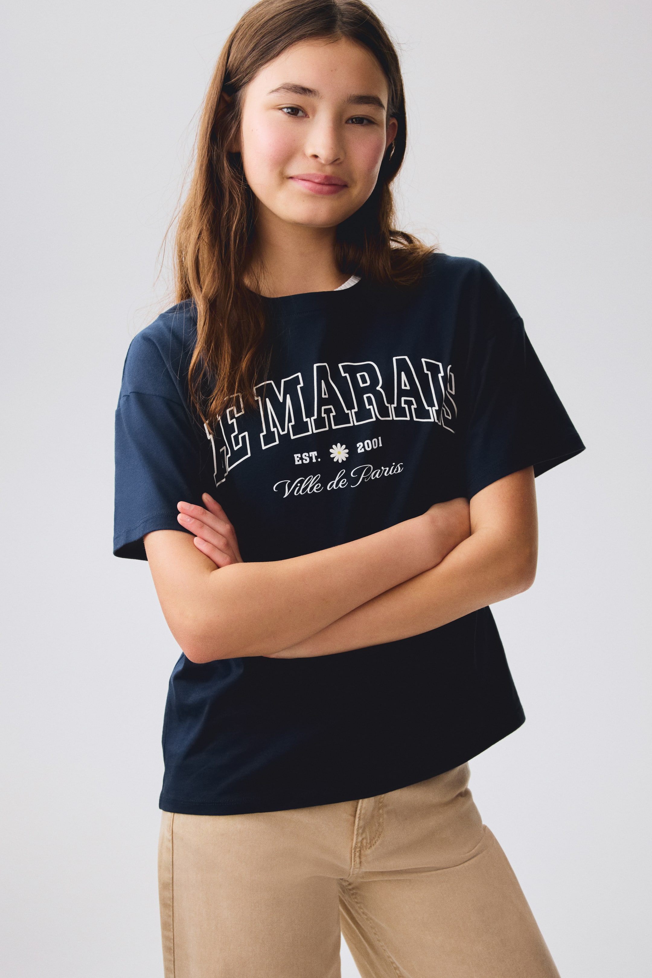 View larger image: A navy blue crew neck t-shirt with short sleeves is showcased, facing forward. It features a white graphic print on the front, reading "LE MARAIS" in outlined block letters, with "EST. 2001" and a small daisy symbol, and "Ville de Paris" in script below.