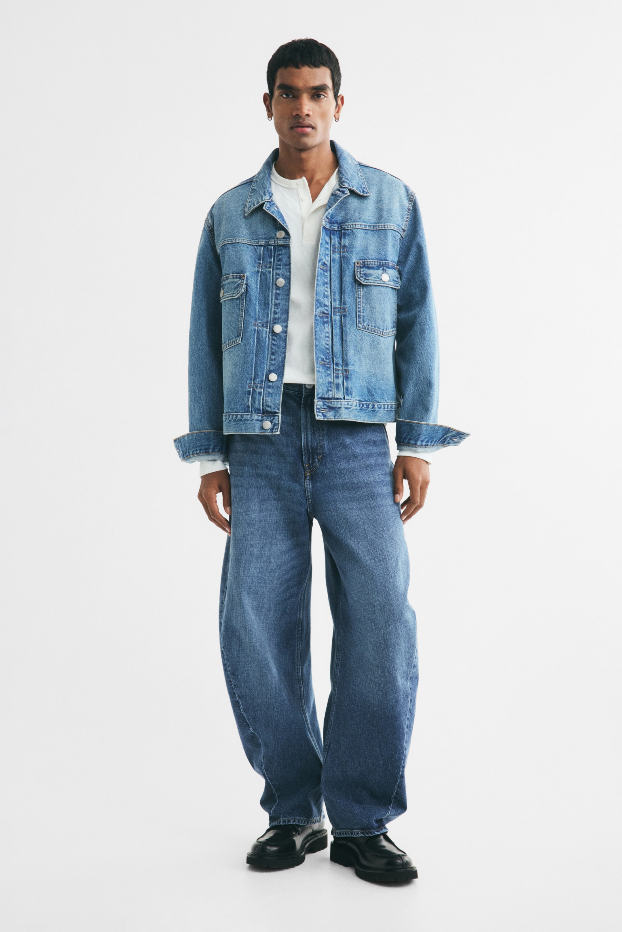 View larger image: The model is wearing a medium-blue denim jacket with a pointed collar and buttoned chest pockets, with the sleeves cuffed and layered over a white Henley shirt. Blue loose-fit, wide-leg jeans are also worn.