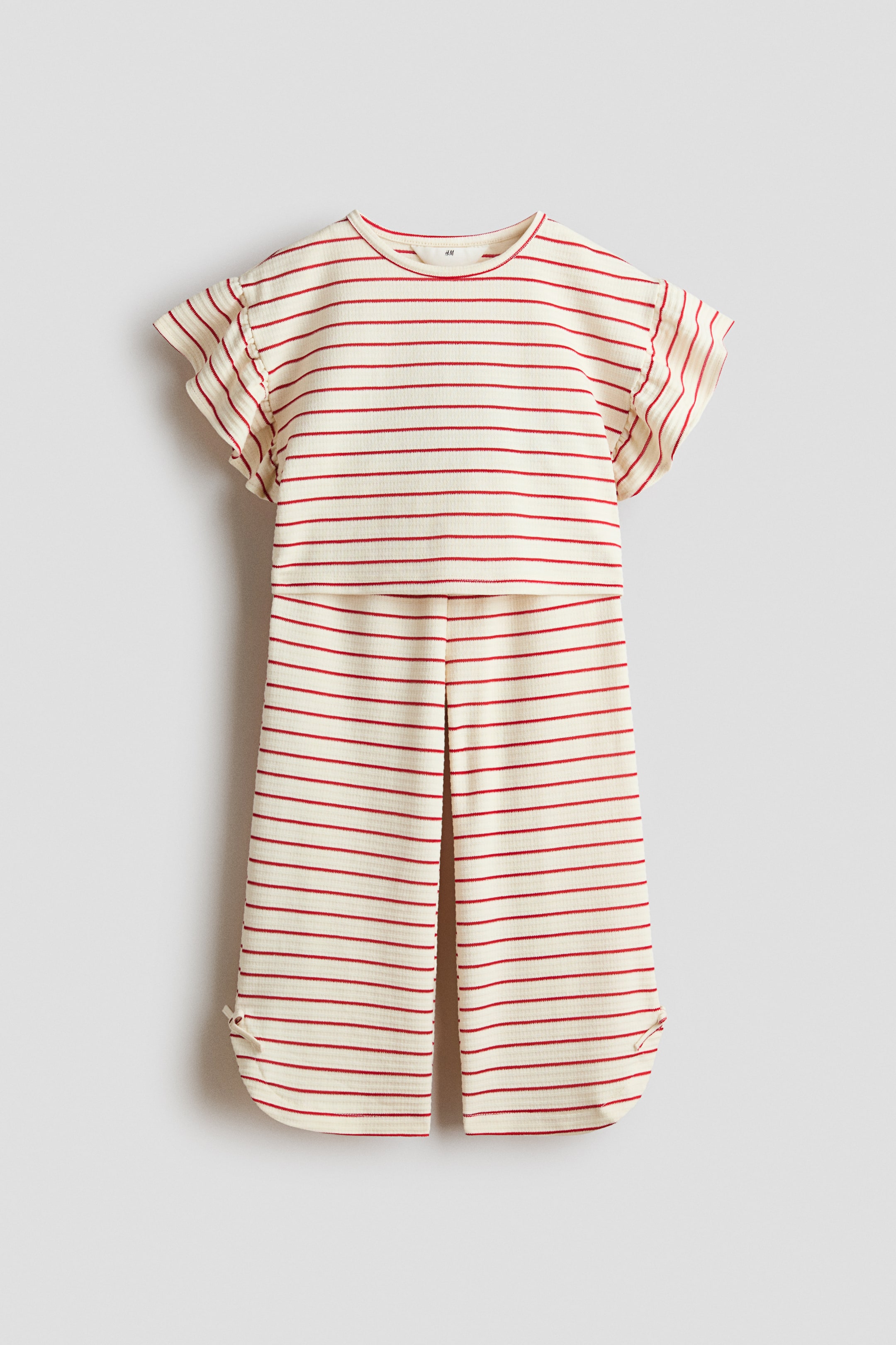 Grösseres Bild ansehen: Vorderansicht eines off-white, gerippten Jumpsuits mit horizontalen, roten Streifen. Er ist mit einem runden Halsausschnitt, kurzen Volantärmeln und einem Cut-out im Taillenbereich gestaltet. Die weit geschnittenen Hosenbeine zeigen eine mittige vordere Falte und geschlitzte Säume, mit einem kleinen Bindeband-Detail am Saum des linken Beins.