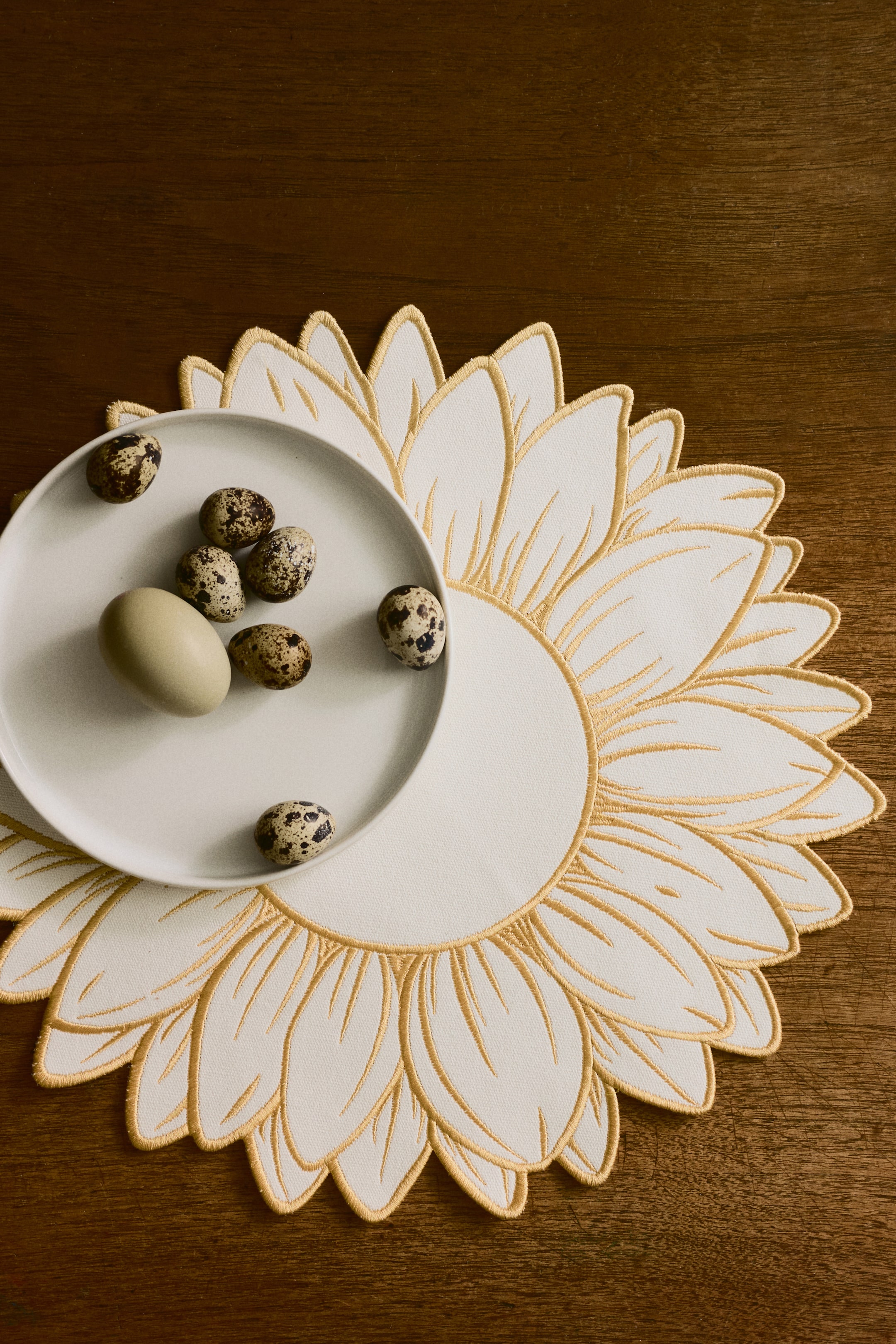 הצגת תמונה גדולה יותר: מפת שולחן לבנה מציגה עיצוב בהשראת חמניות. פרטי רקמת סאטן זהובים יוצרים את קווי המתאר והוורידים של עלי הכותרת, וצלחת לבנה מונחת במרכז המעגלי של מפת השולחן.