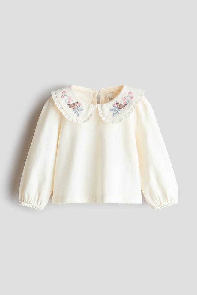 An off-white cotton jersey blouse is facing front, showcasing a rounded collar with frill-trimmed lace edges and small embroidered bird and floral details. The blouse features long balloon sleeves with narrow elasticated cuffs. - 1