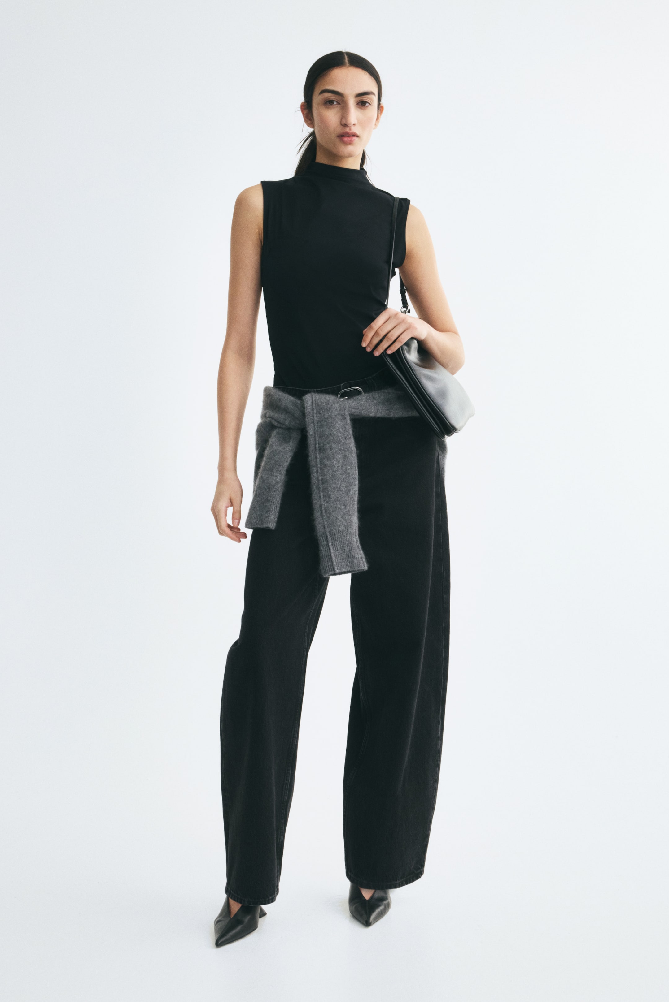 Voir une image plus grande: Un haut noir sans manches à col montant et un jean noir à jambes larges sont portés par le mannequin, agencés avec un chandail en maille gris noué à la taille. Un sac à bandoulière en cuir noir est porté à l’épaule, complétant le look.