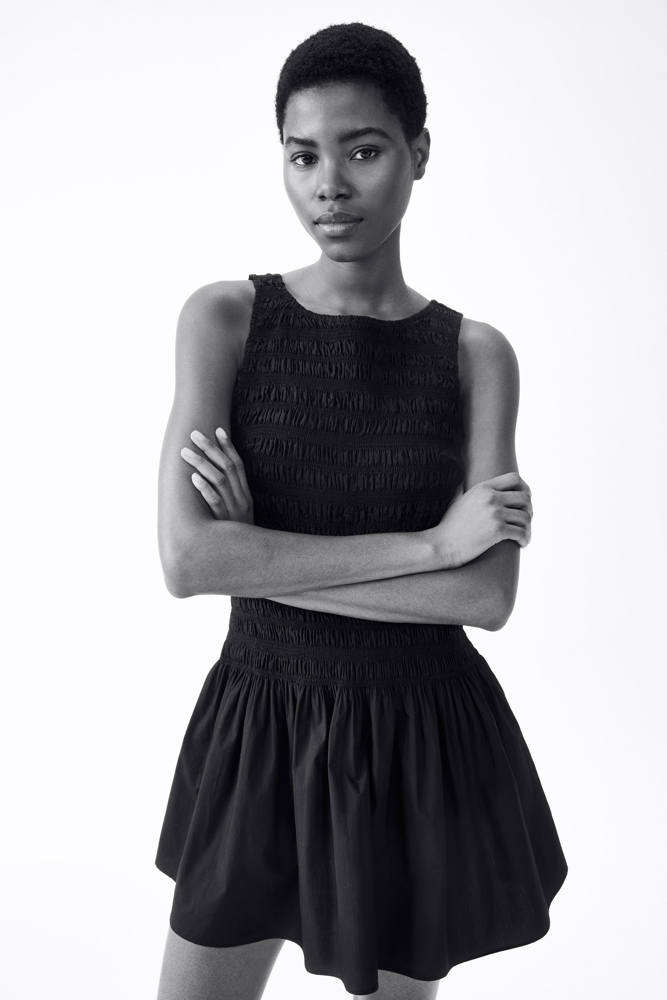 View larger image: The model is donning a short, sleeveless dress crafted from a cotton weave. It showcases a smocked top section, a round neckline, and a flared skirt. The model stands with arms crossed.