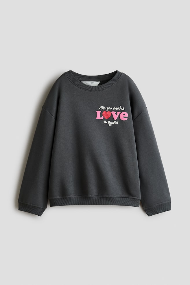 Una felpa oversize grigio scuro è mostrata frontalmente, con scollo tondo a coste, spalle scese e maniche lunghe con polsini a coste. Sul petto è presente una stampa grafica con la scritta "All you need is LOVE" in rosa e bianco con un cuore rosso, e sotto "The Beatles". - 1