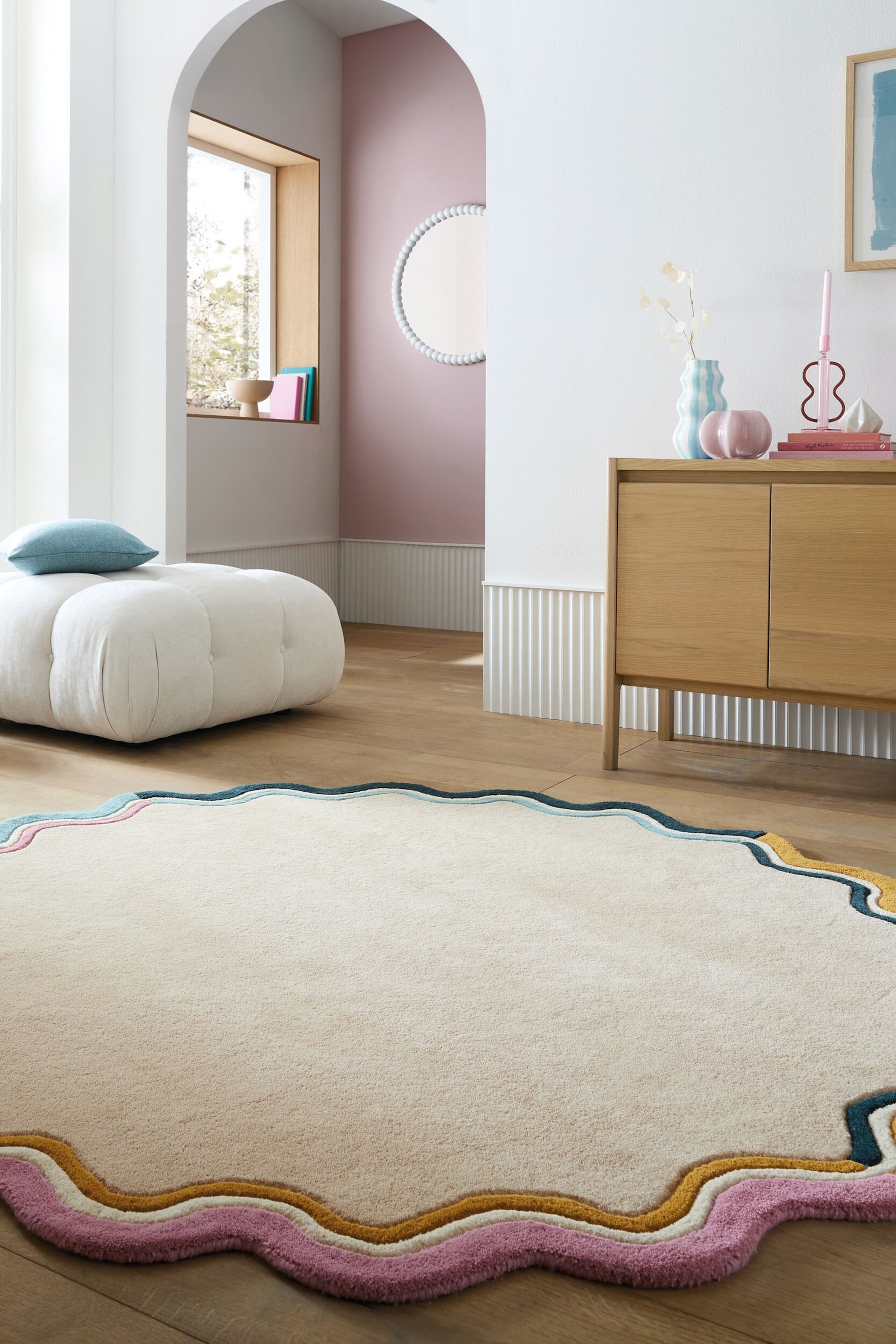 Agrandir l'image: Un tapis présente un contour irrégulier et ondulé. Son corps principal est de couleur blanc cassé, et sa bordure est détaillée avec des bandes ondulées superposées en rose, rose clair, jaune et turquoise.