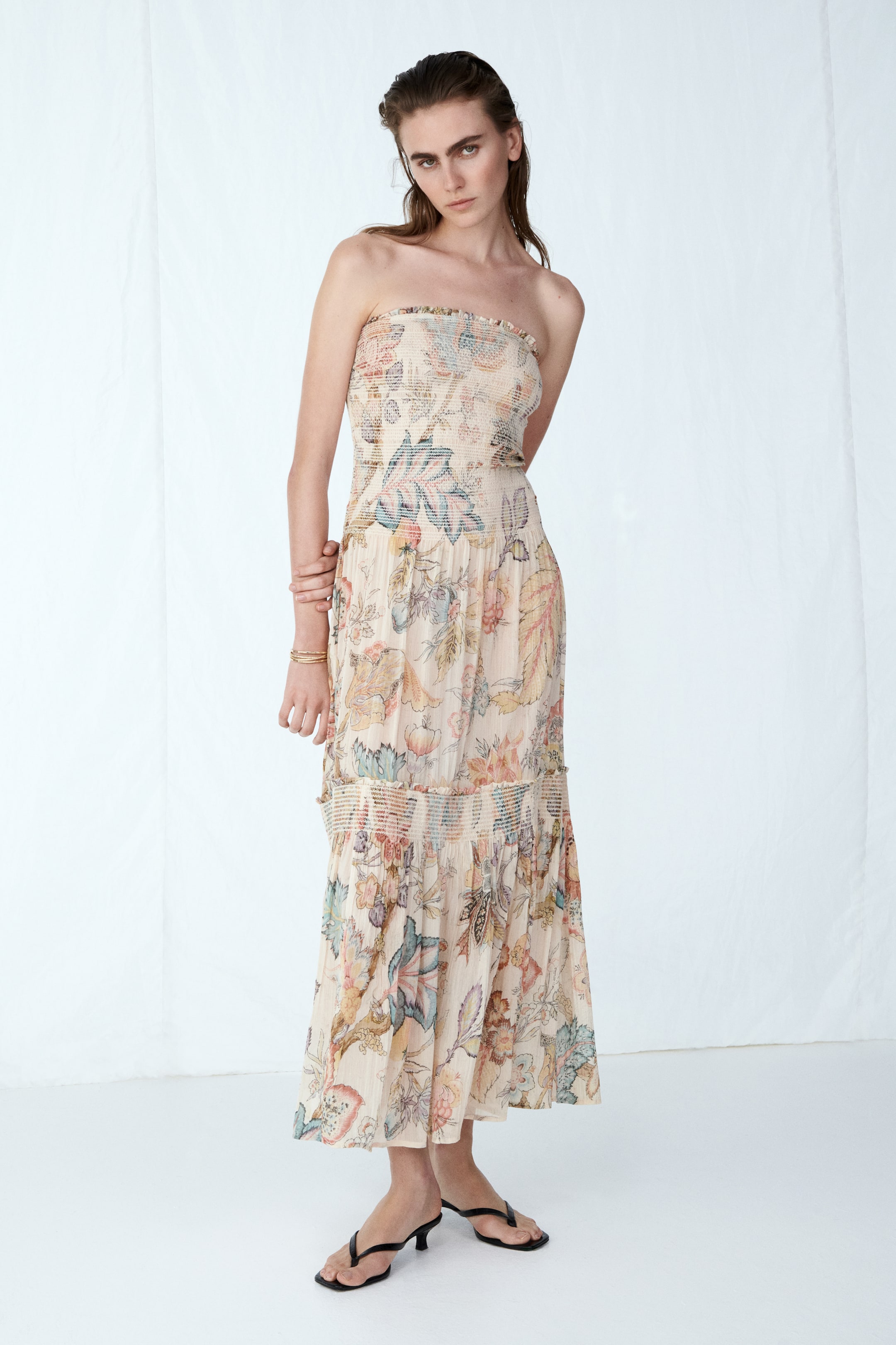View larger image: A strapless dress is displayed from the front, showcasing a beige backdrop adorned with a vibrant floral and leaf motif. It is crafted with a smocked bodice and a tiered skirt, featuring a smocked band detail below the waist.
