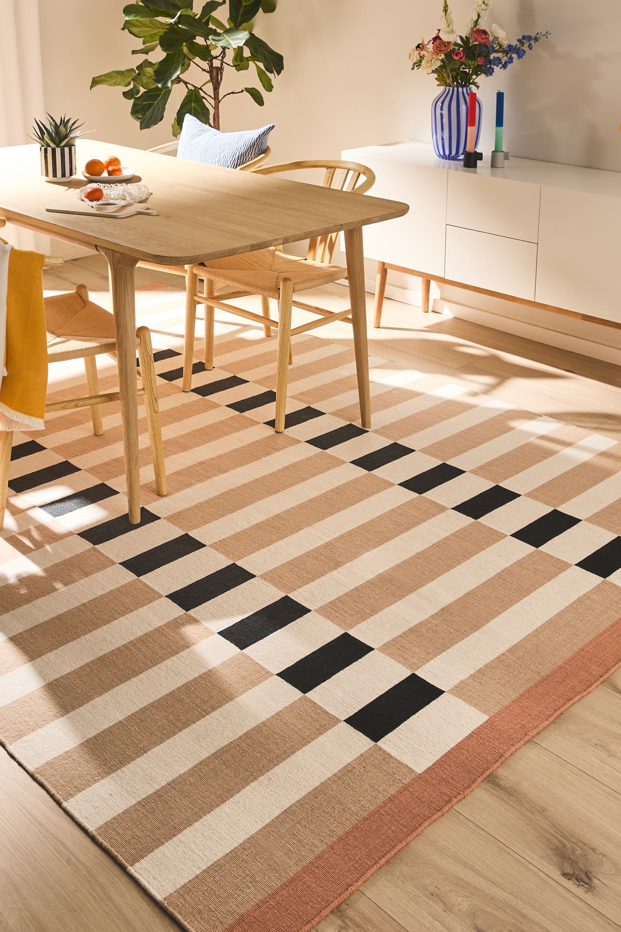 Ver imagen más grande: Una alfombra rectangular presenta franjas horizontales en crema, beige y negro. Las franjas negras forman un patrón de tablero de ajedrez escalonado, y un borde sólido de color marrón rojizo recorre un borde visible.