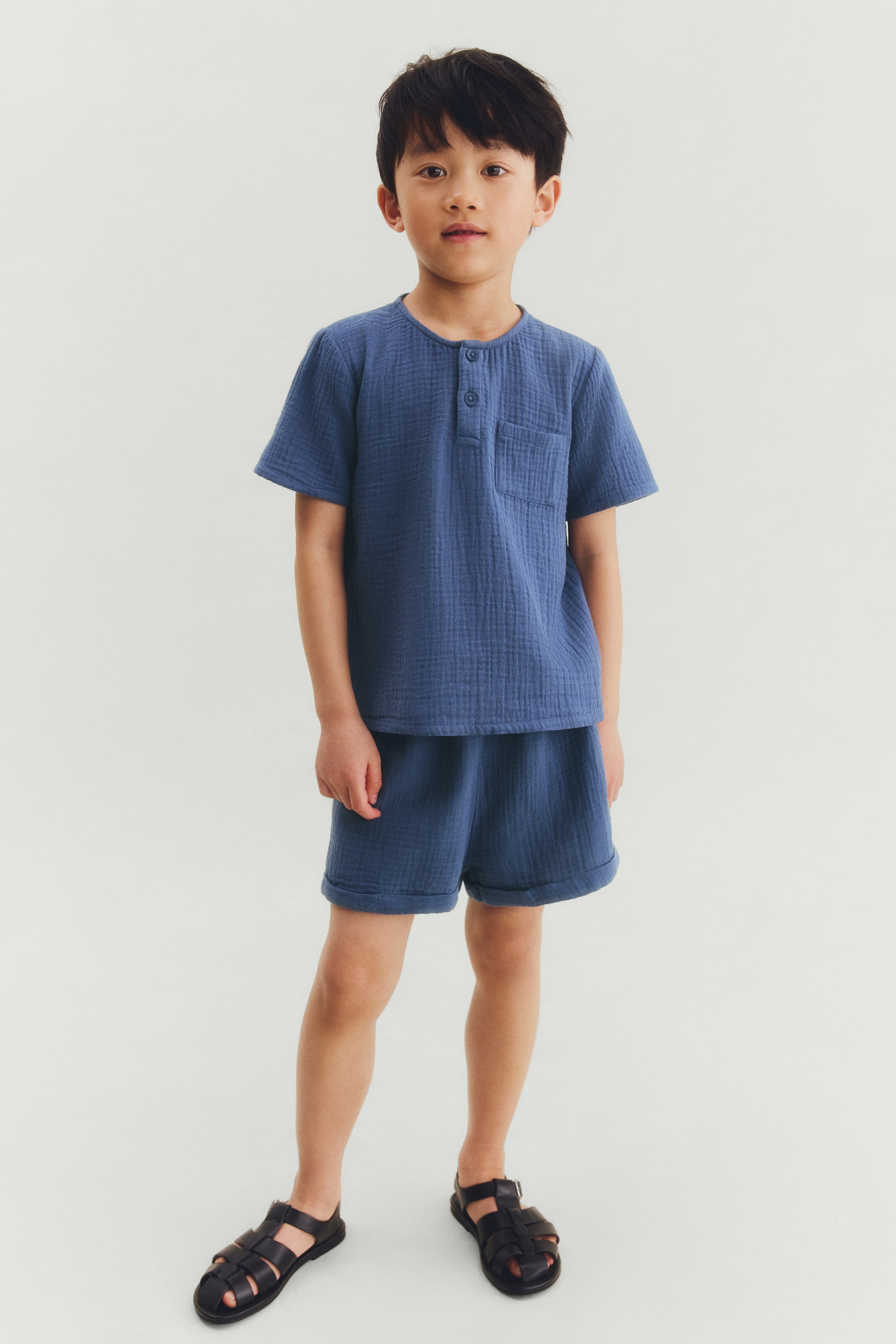 Grotere afbeelding bekijken: Een blauw tweedelig setje wordt frontaal gepresenteerd, bestaande uit een top met korte mouwen en bijpassende shorts. De top heeft een Henley-halslijn met twee knopen en een borstzak, terwijl de shorts omgeslagen zomen hebben. De gehele set is gemaakt van een getextureerde, gekreukelde stof.