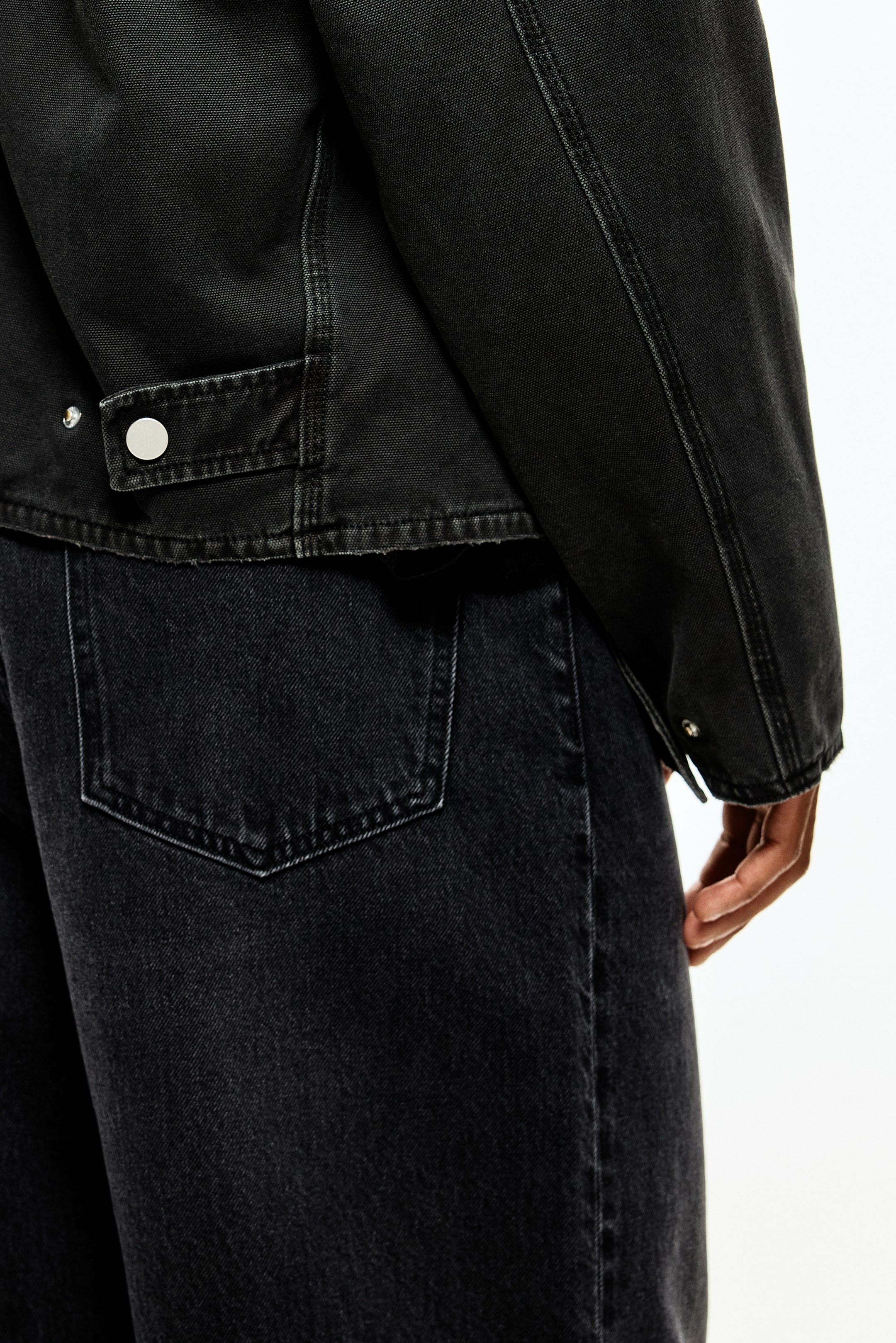 Ampliar la imagen: Una chaqueta de estilo de ropa de trabajo de color gris oscuro se lleva sobre unos jeans de mezclilla gris oscuro, vista desde un ángulo lateral y trasero. La chaqueta está mostrando una lengüeta con un broche de botón plateado en el borde y costuras visibles a lo largo de la manga, mientras que los jeans muestran un bolsillo trasero parche.