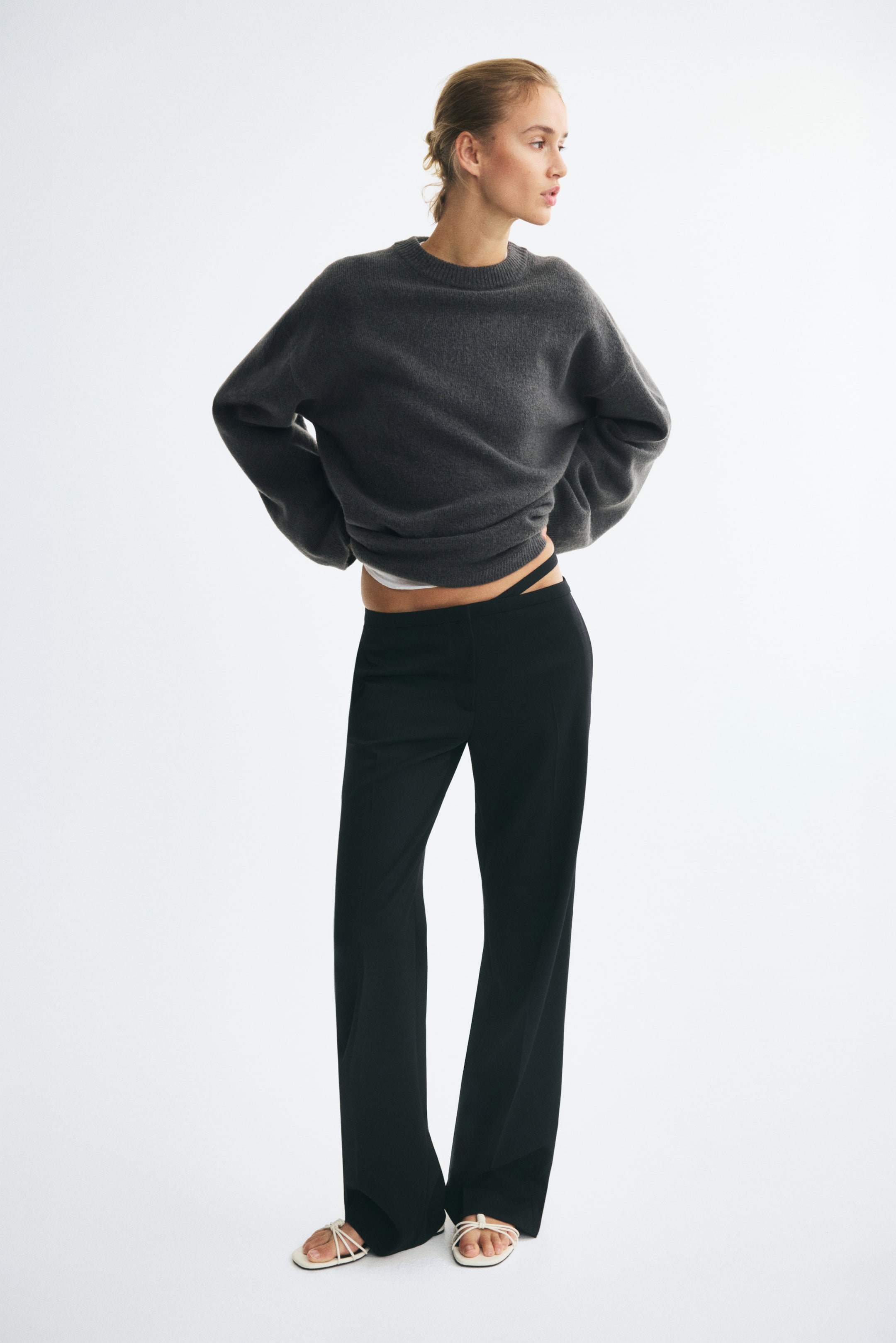 View larger image: An oversized dark gray knit sweater featuring a ribbed crew neck, dropped shoulders, and long sleeves is worn by the model. The model is posing with their hands resting on their hips, pairing the sweater with black wide-leg trousers and white strappy sandals.
