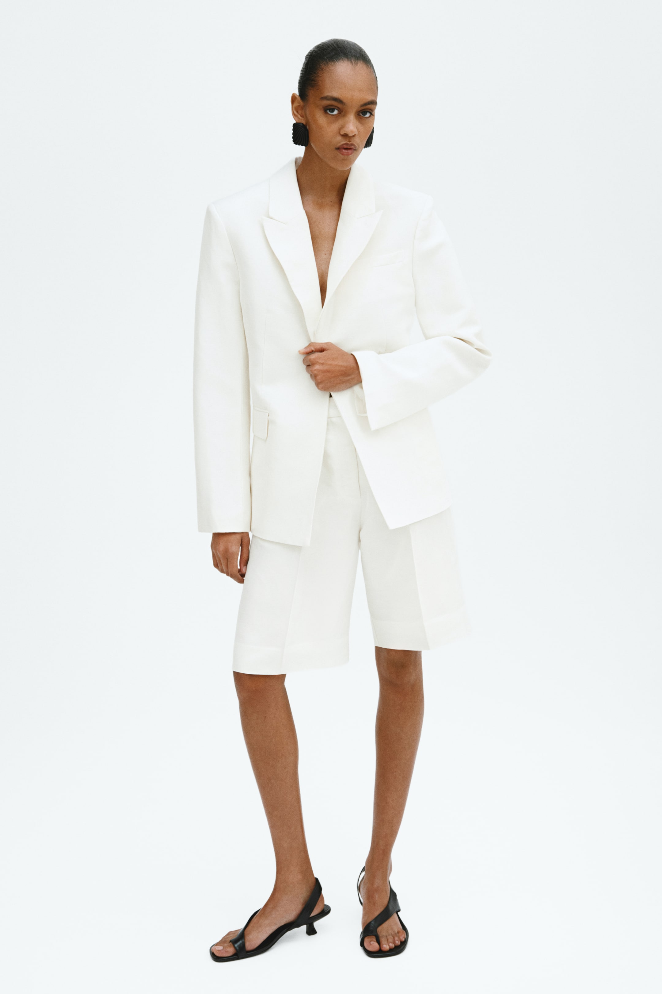 Ver imagem maior: Um blazer branco e calções a condizer, de comprimento pelo joelho, estão a ser exibidos de um ângulo frontal. O blazer apresenta lapelas em bico, ombros estruturados, mangas compridas e um bolso com aba, enquanto os calções ajustados completam o conjunto coordenado.