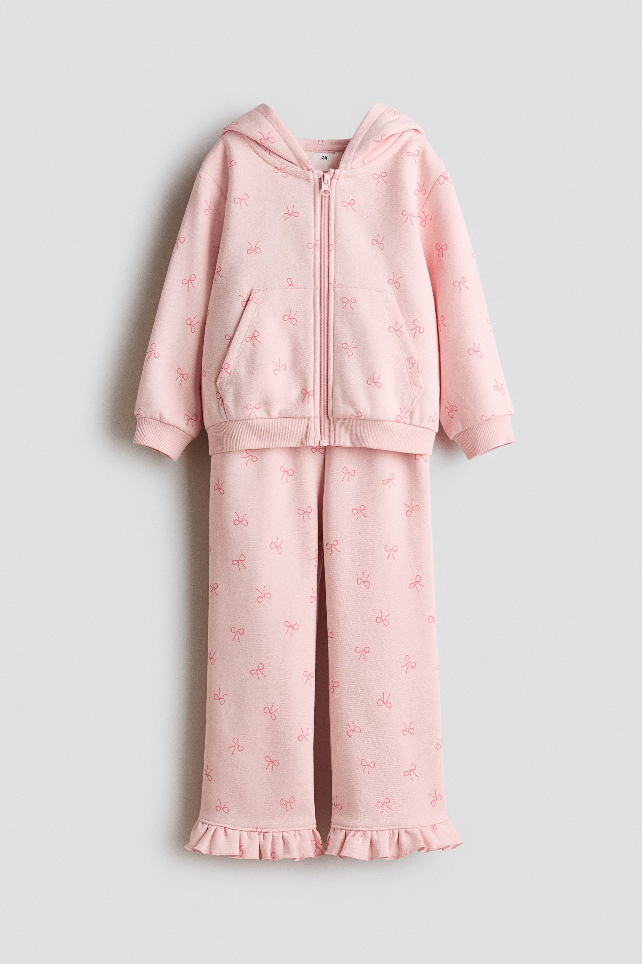 View larger image: A pink two-piece tracksuit is hanging, featuring a full-zip hoodie and matching pants, both patterned with repeating darker pink bow outlines. The hoodie includes a hood, a kangaroo pocket, and ribbed cuffs and hem, while the pants are showcasing ruffled hems at the ankles.