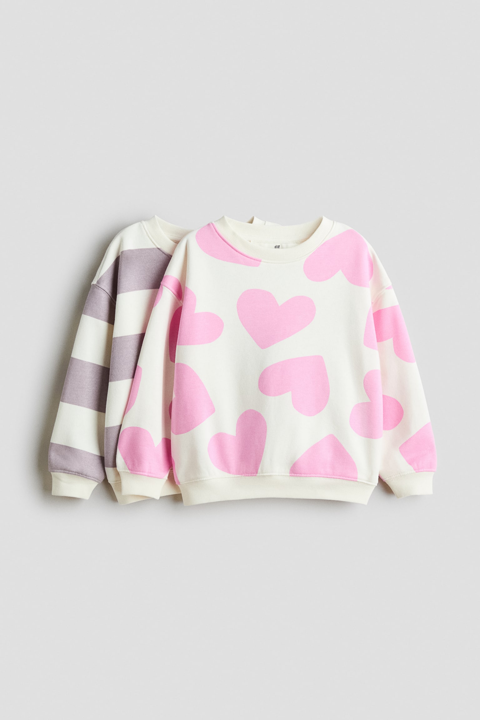 Two long-sleeved sweatshirts are shown in a front-facing view, with one layered slightly behind the other. The front sweatshirt features an all-over pink heart pattern on an off-white base, while the rear sweatshirt shows wide, horizontal purple-grey stripes on a matching off-white base. Both garments feature ribbed crew necks, cuffs, and hems.